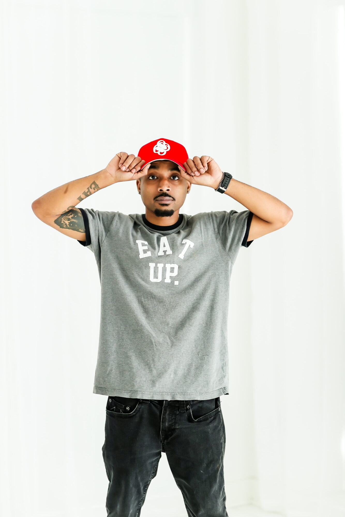Young man wearing a gray T-shirt with text, black pants, and a red cap, standing against a white background, adjusting his cap.