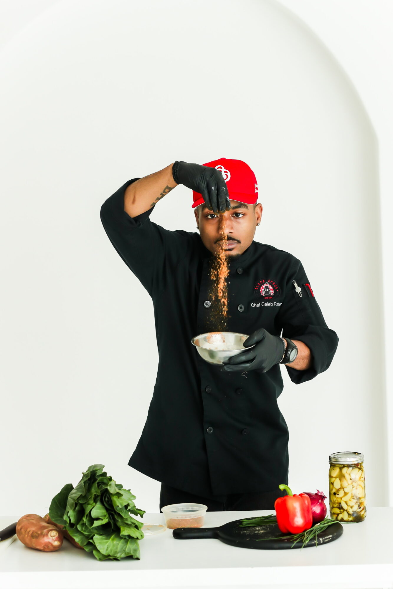 Chef in black uniform and red bandana sprinkling spices into a bowl, with vegetables on a white background.