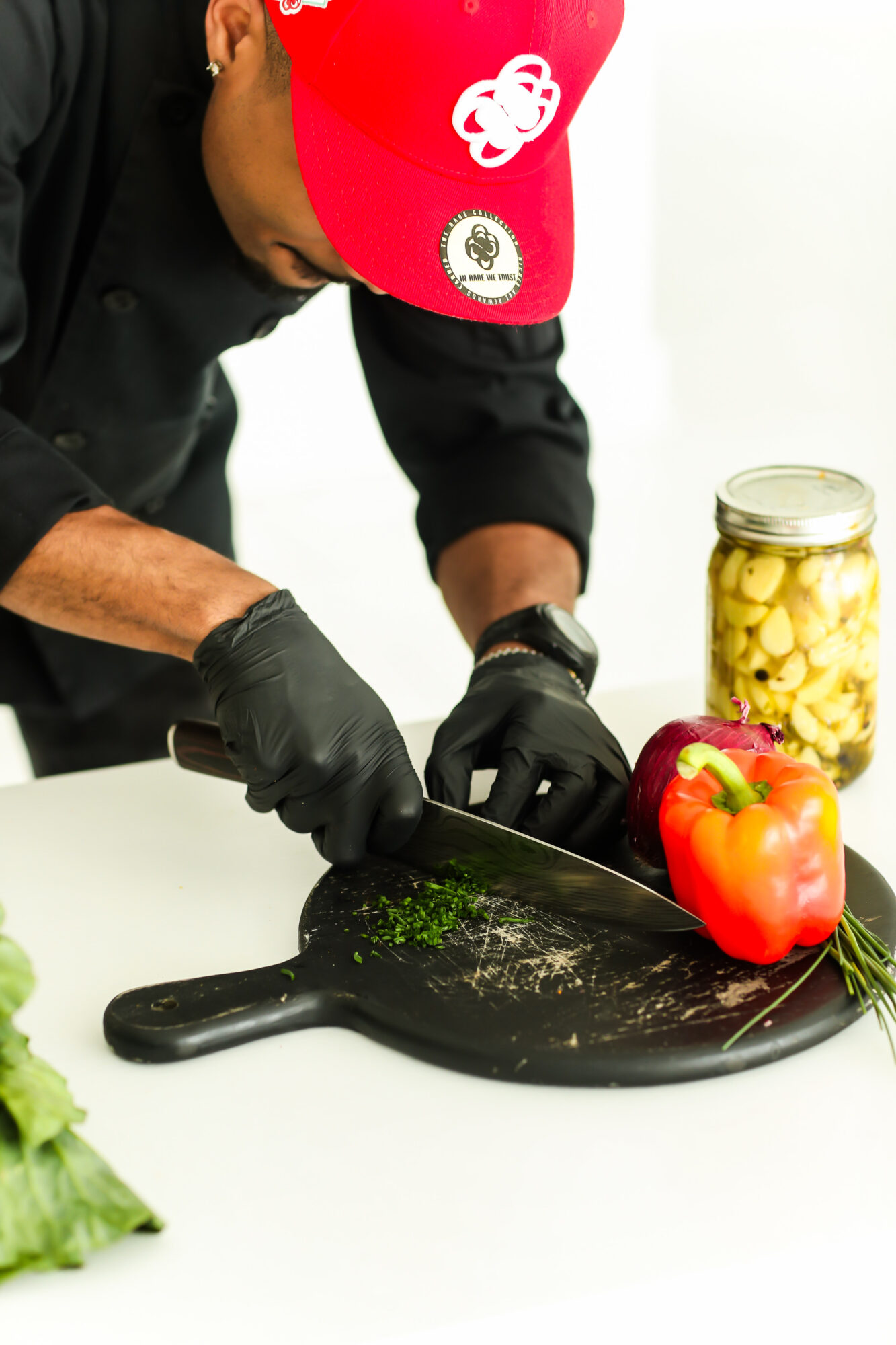 Person chopping herbs on a black cutting board with a knife, wearing a red cap and black gloves, with a jar of yellow pickles and a red bell pepper nearby.