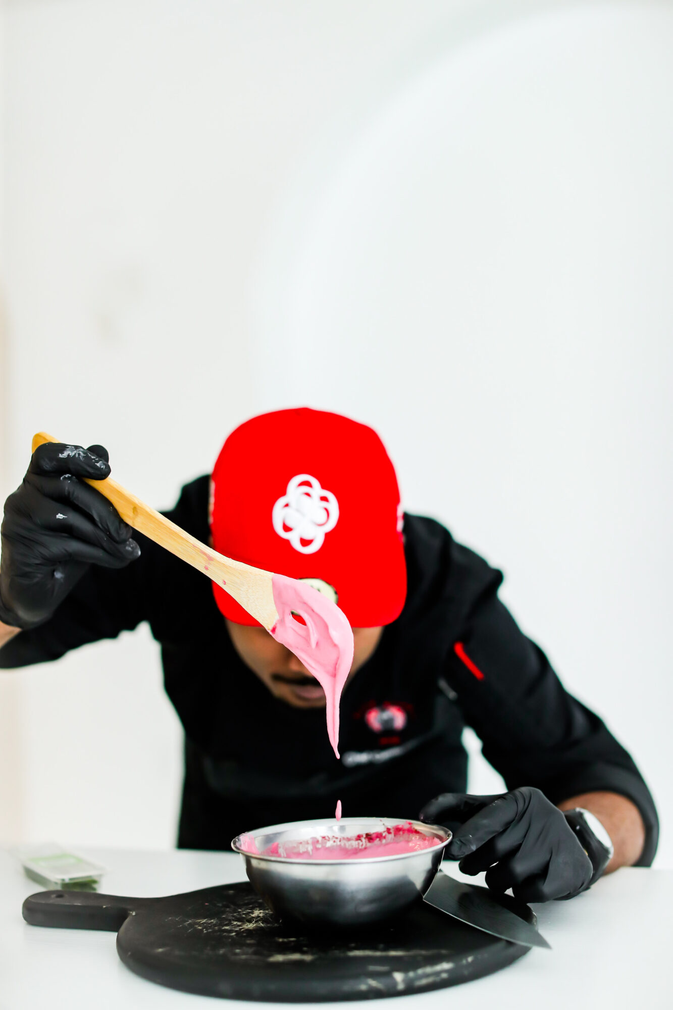 Person wearing a red cap and black gloves pouring pink liquid into a bowl, with a spoon in hand.