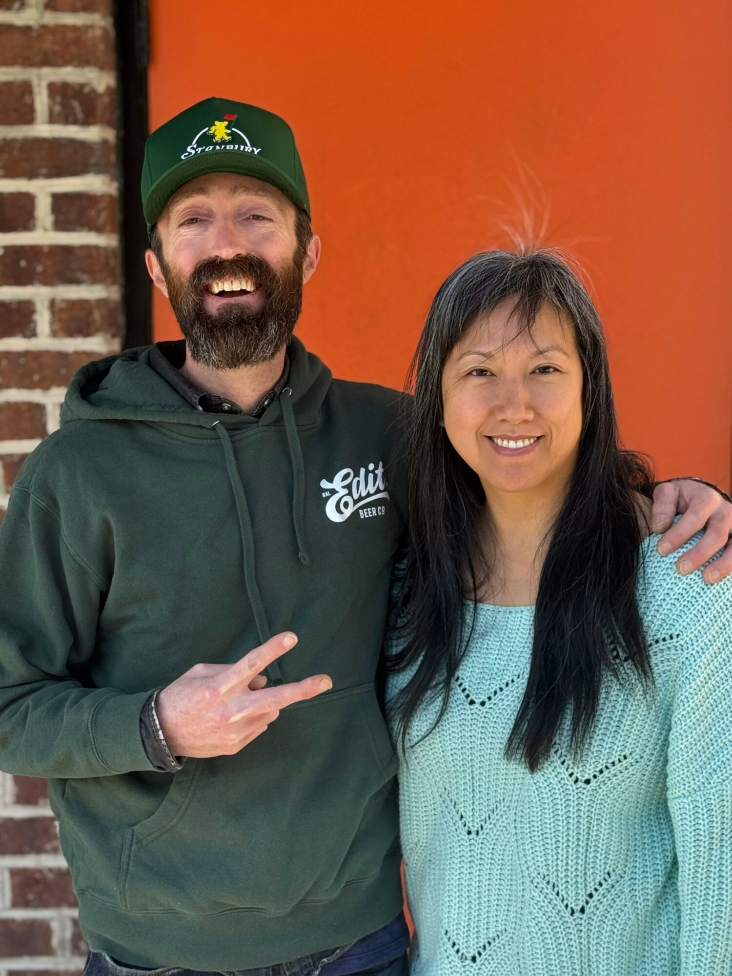 A smiling man with a beard and a woman with long dark hair stand together outdoors, with a brick wall and orange background behind them.