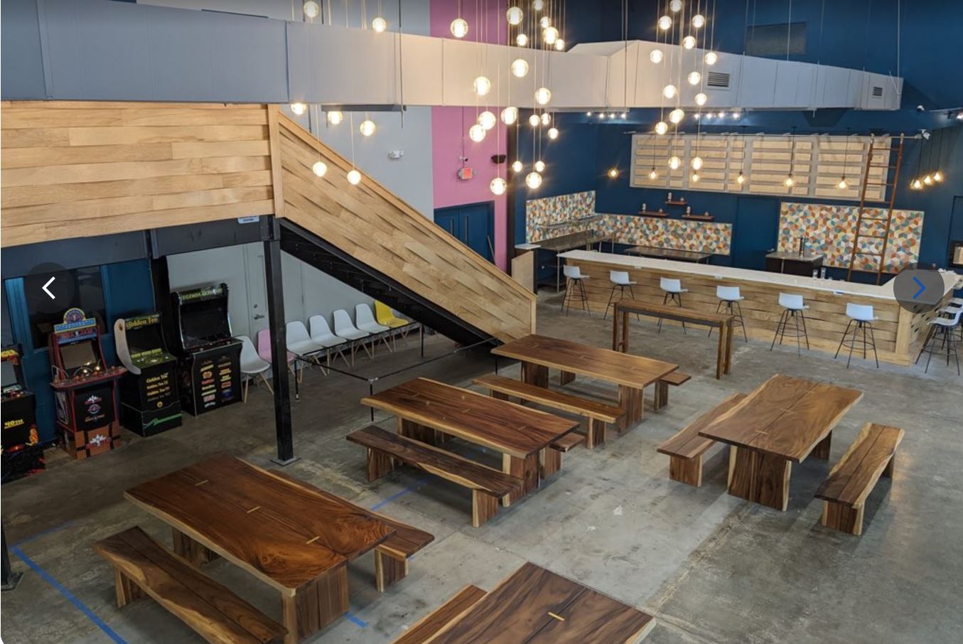 Interior of a modern restaurant with wooden tables, benches, and a staircase, illuminated by hanging lights.
