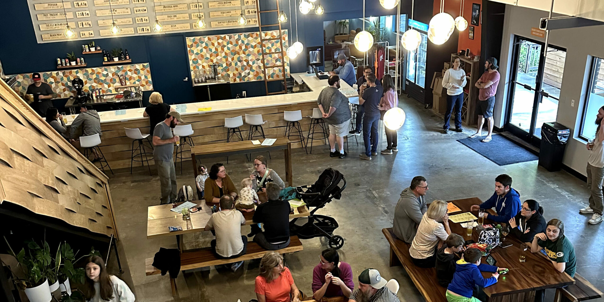 People gather in a modern cafe with seating, a bar, and large windows, some standing and some sitting at tables.