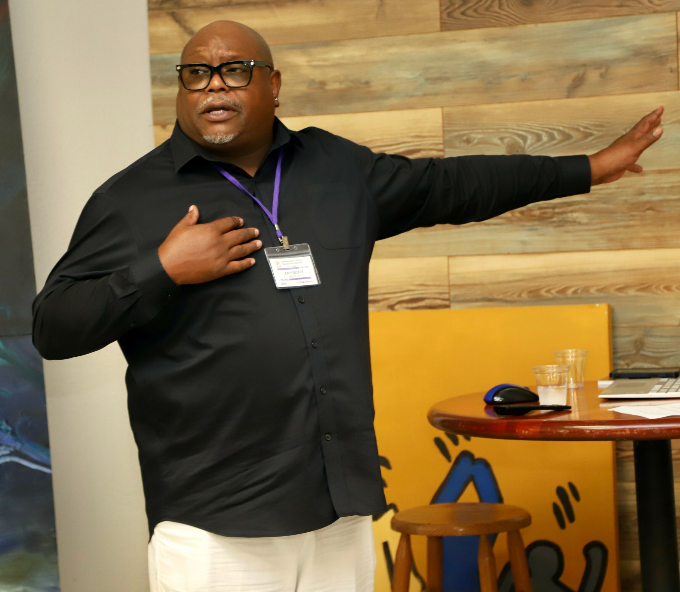 Man in black shirt and glasses gestures with arm extended in a room with wooden wall and yellow furniture.