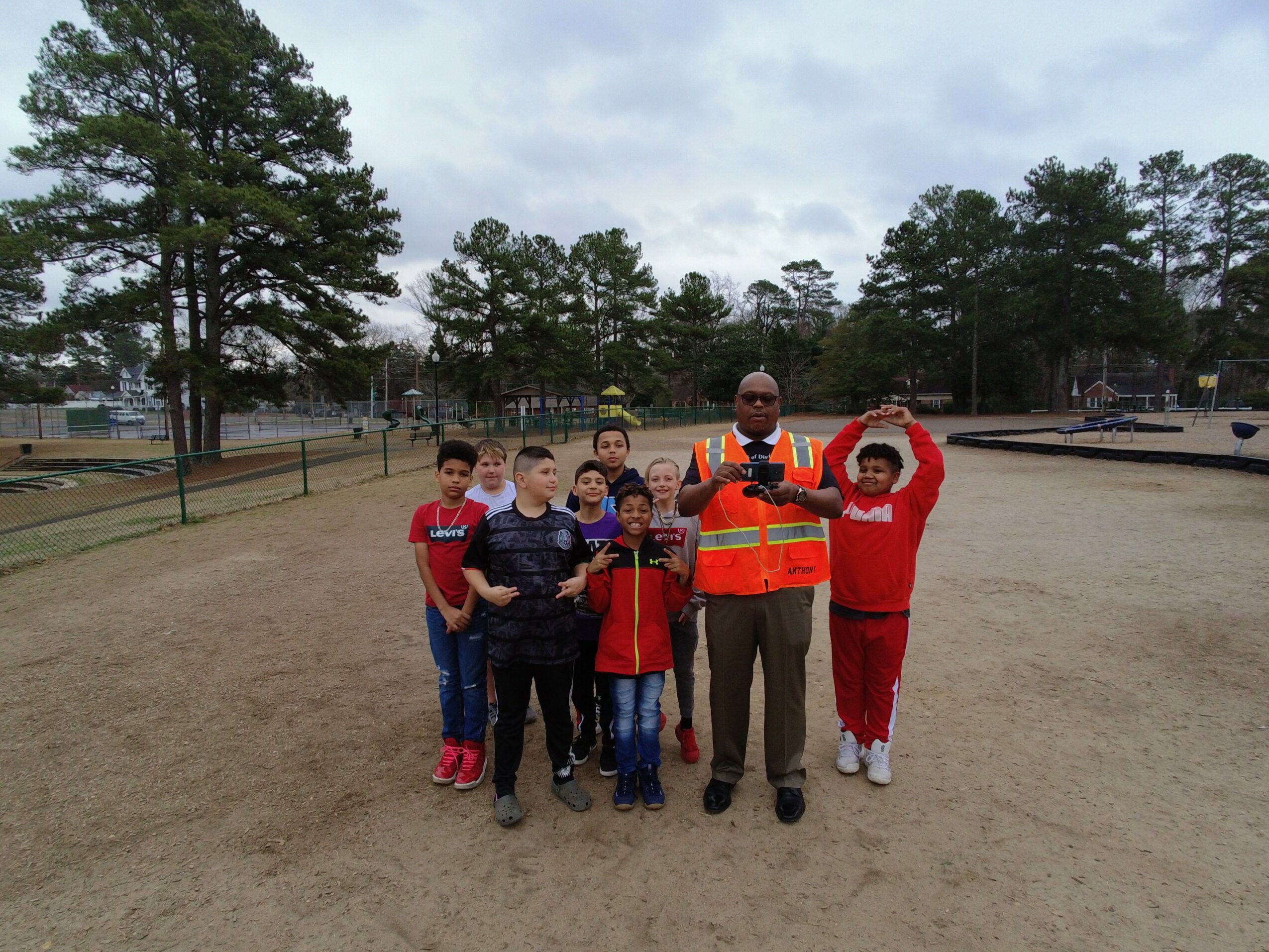 Group of children and an adult standing outdoors on a dirt field, some children wearing red shirts, trees in background.
