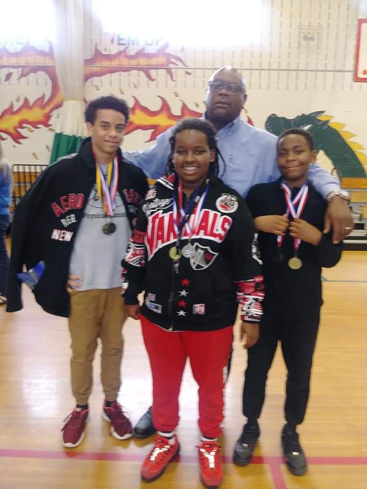Four people standing in a gymnasium, three children with medals and an adult man behind them, colorful wall decorations in the background.