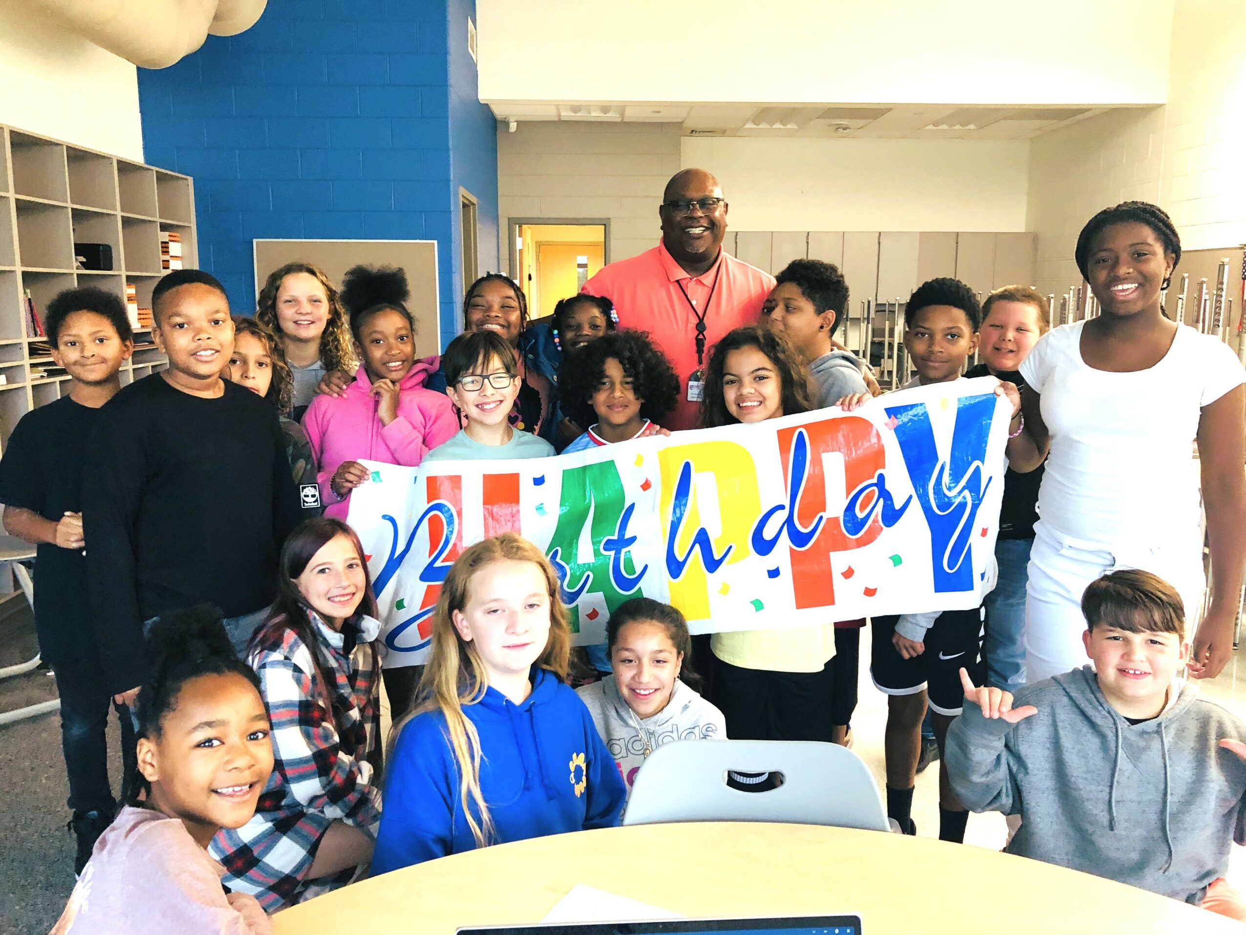 Group of children and an adult holding a colorful birthday banner in a classroom, smiling and celebrating.
