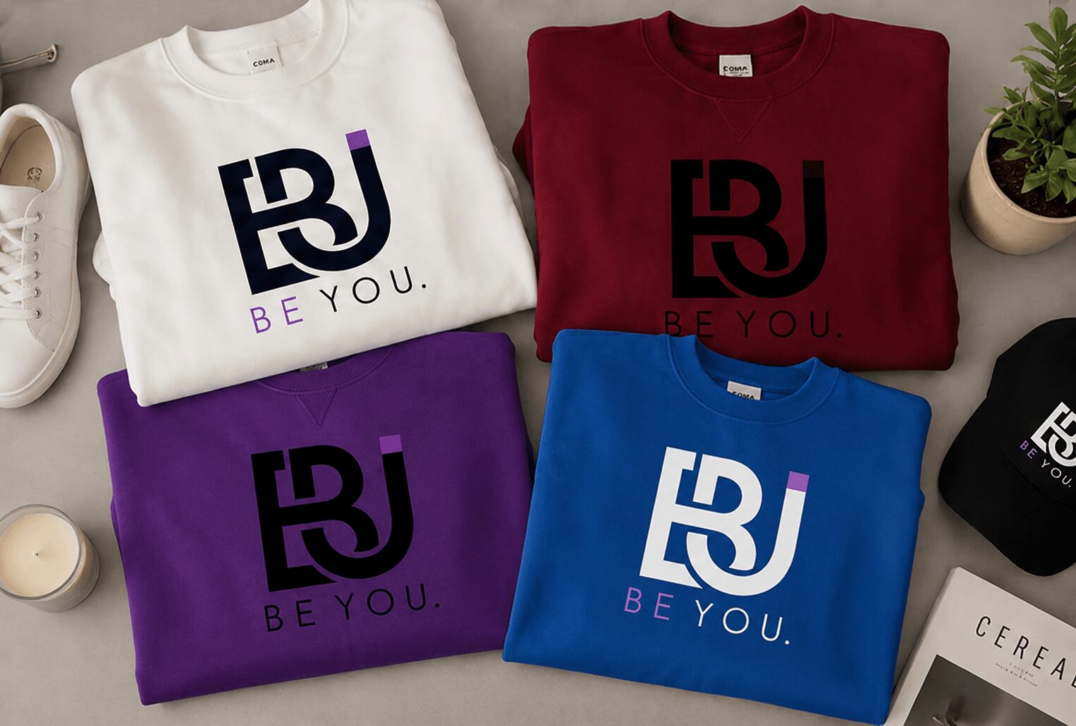 Four folded sweatshirts with bold logo and text, in white, red, purple, and blue, arranged on a surface with plants and books.