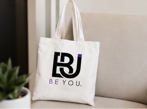 White tote bag with black and purple logo and text, placed on a beige sofa.