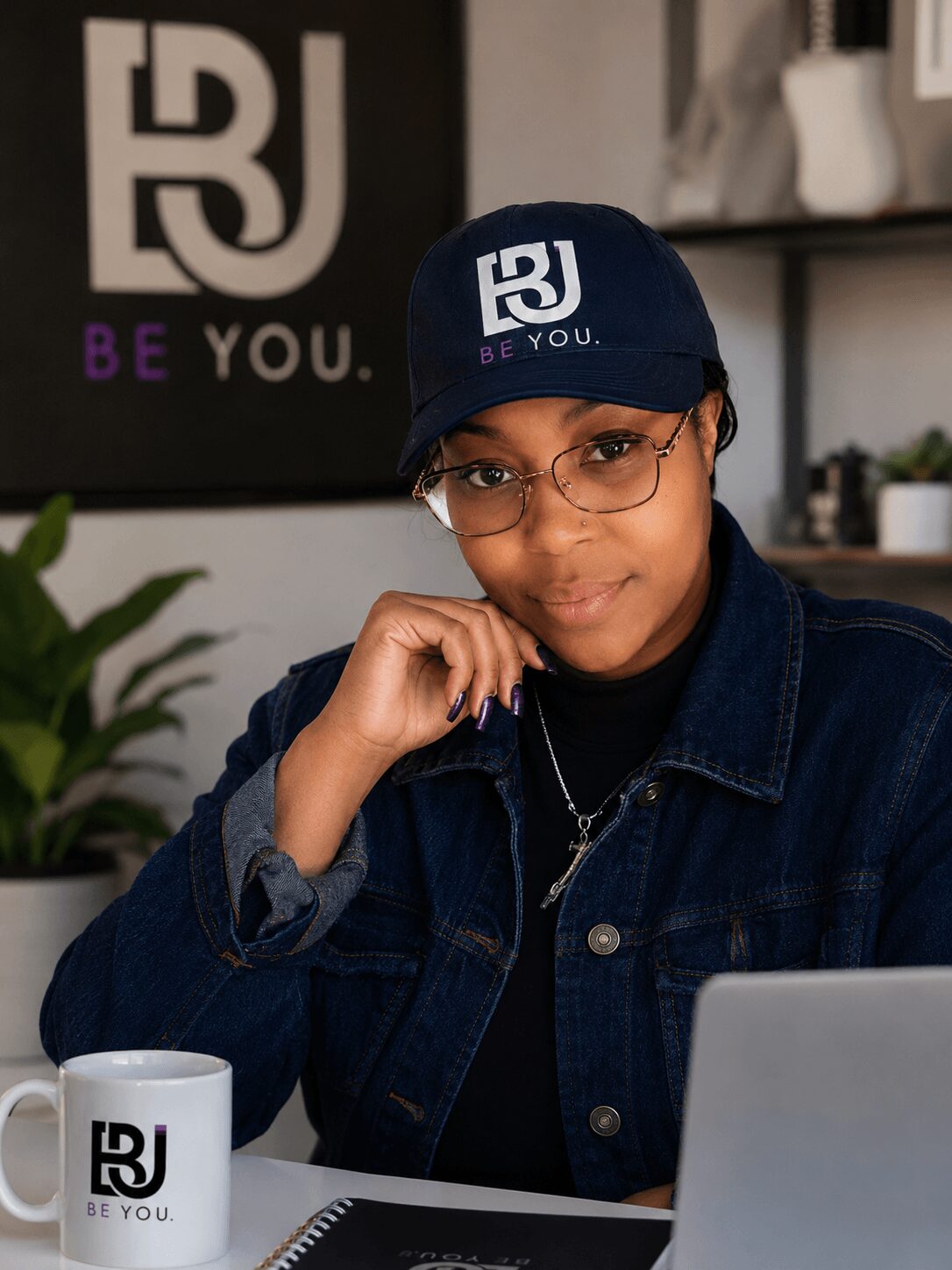 Young person wearing glasses and a cap with 'BJ' logo, sitting at a desk with a mug and laptop, smiling.