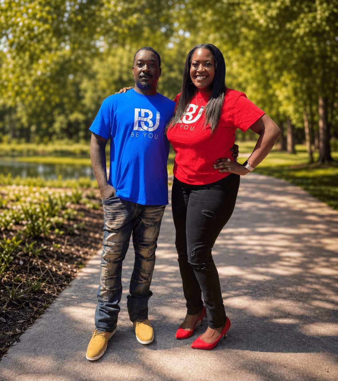 Two people standing on a park path, smiling, with trees and greenery in the background.