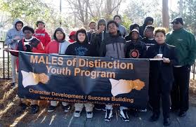 Group of young people holding a banner outdoors, trees in background, sunlight, some wearing hoodies and jackets.