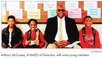 Man in sunglasses and suit sitting with three young students, all smiling, in front of colorful posters, group photo.