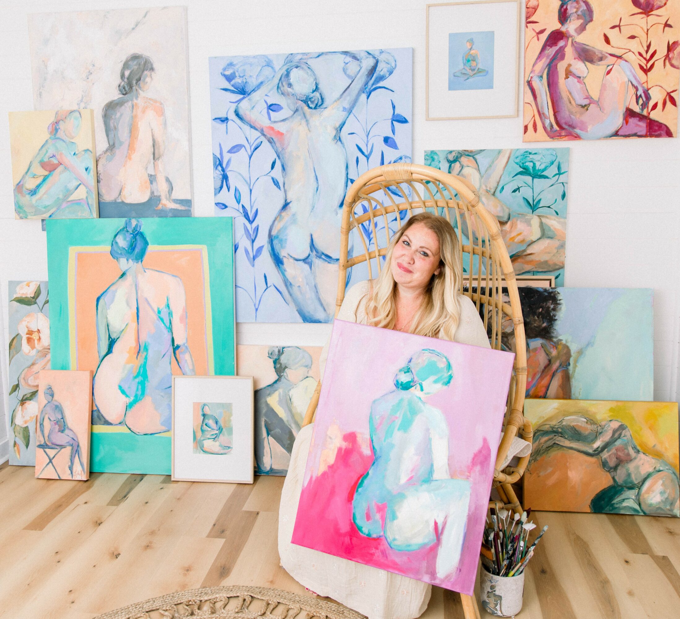 A woman with blonde hair sitting in a wicker chair surrounded by colorful paintings of female figures, some on the wall and others on the floor.