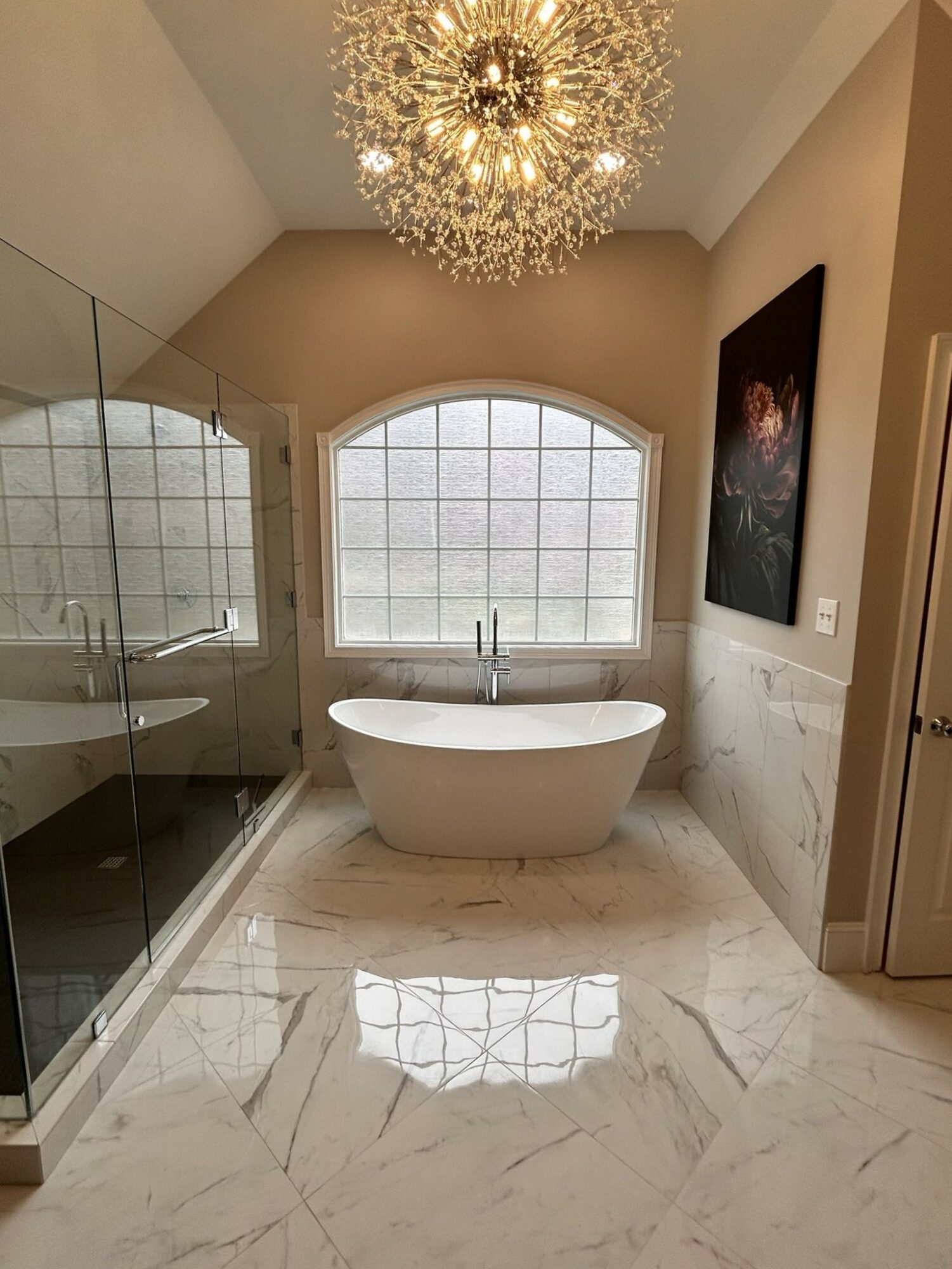 Bathroom with a freestanding bathtub in front of a large arched window, marble floor, glass shower, and chandelier.