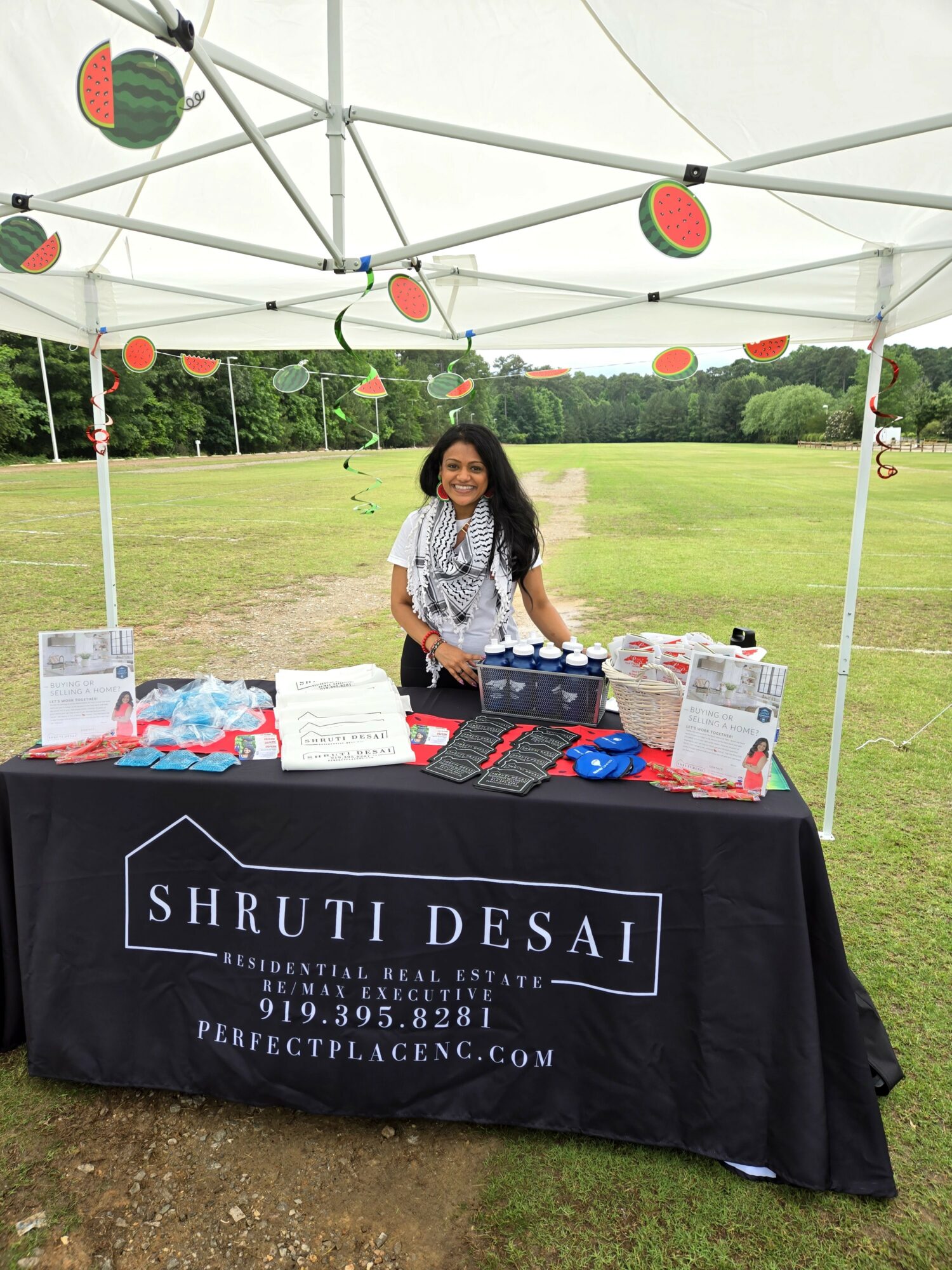 Young girl standing behind a table under a tent with watermelon decorations, outdoors in a grassy area, promotional materials on table.
