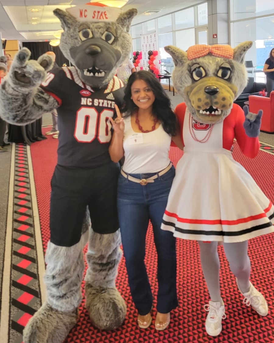 Woman standing between two people in animal costumes inside a building with large windows, red carpet, and chairs.