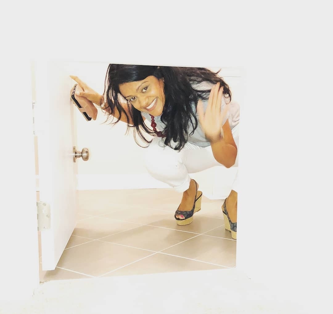 Person crouching and peeking through a small door, smiling, with dark hair and wearing high heels.