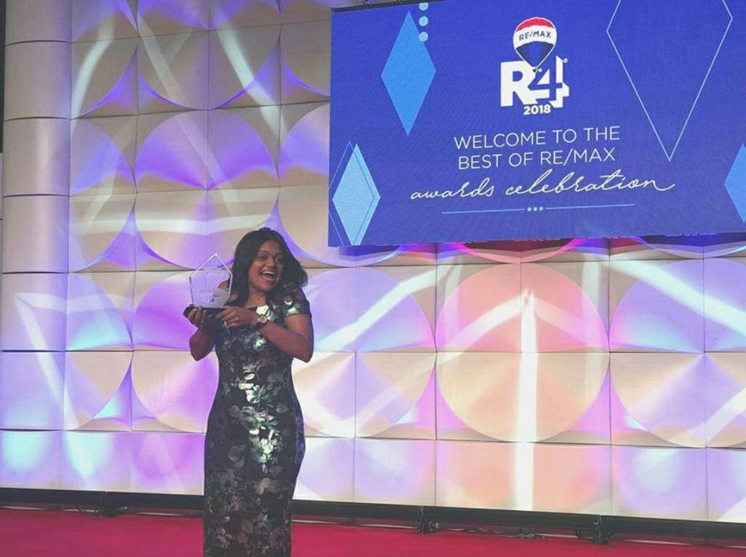 Woman in a black dress standing on stage with a colorful backdrop and a large screen displaying event information.