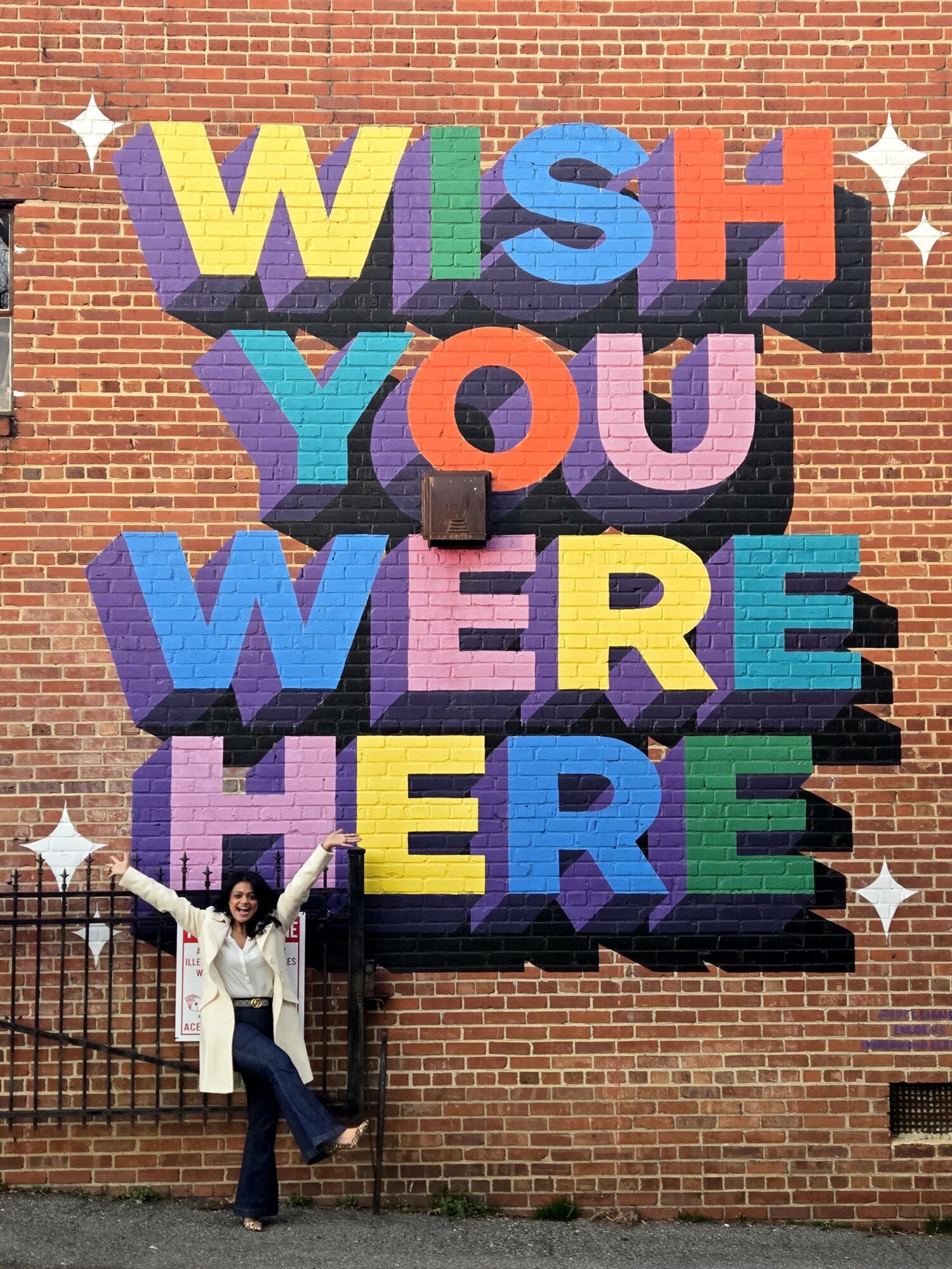 Person standing in front of a brick wall with colorful 3D letters spelling 'WISH YOU WERE HERE', arms raised.