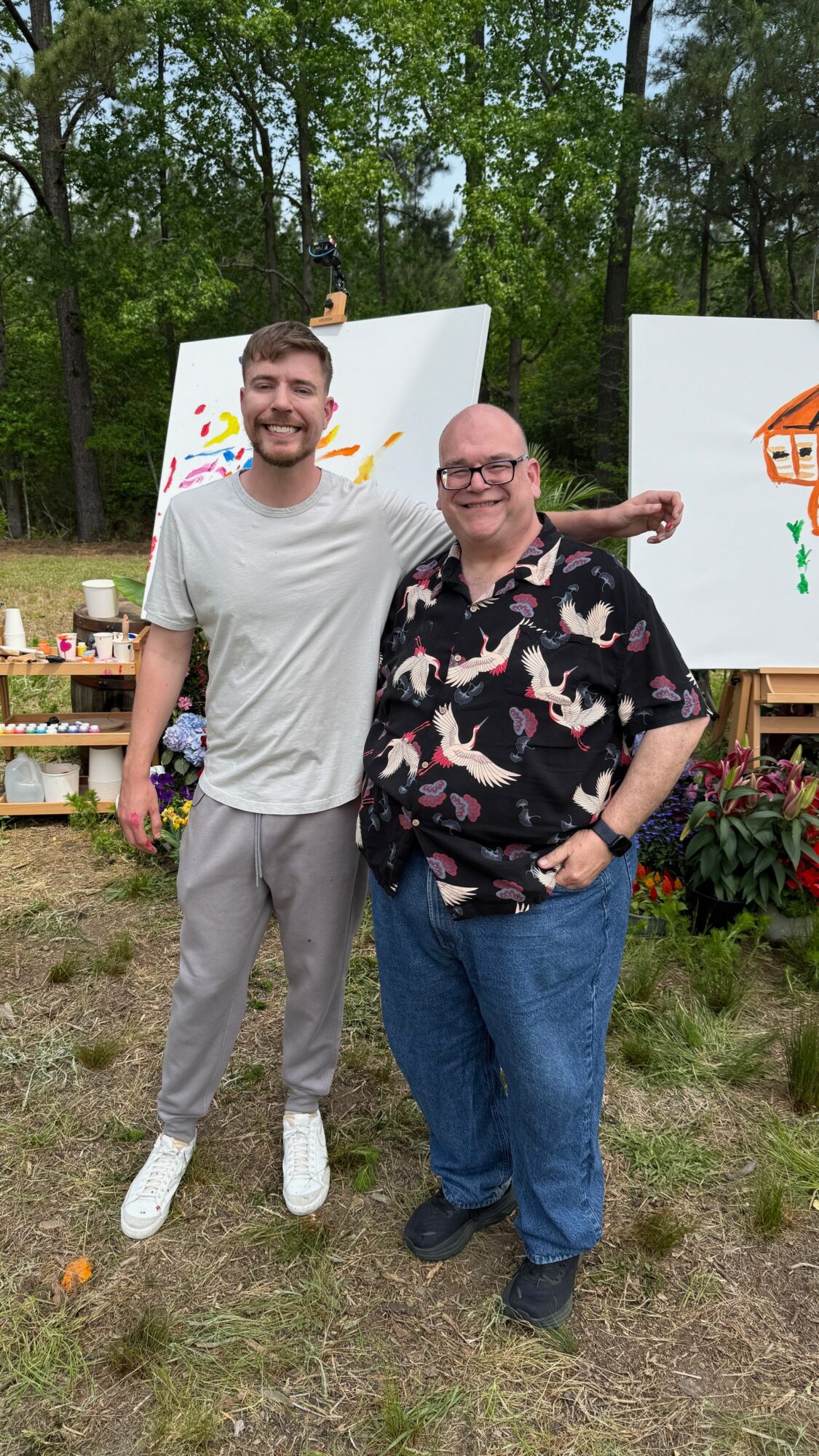 Two men standing outdoors in front of paintings on easels, smiling, with trees and flowers in the background.