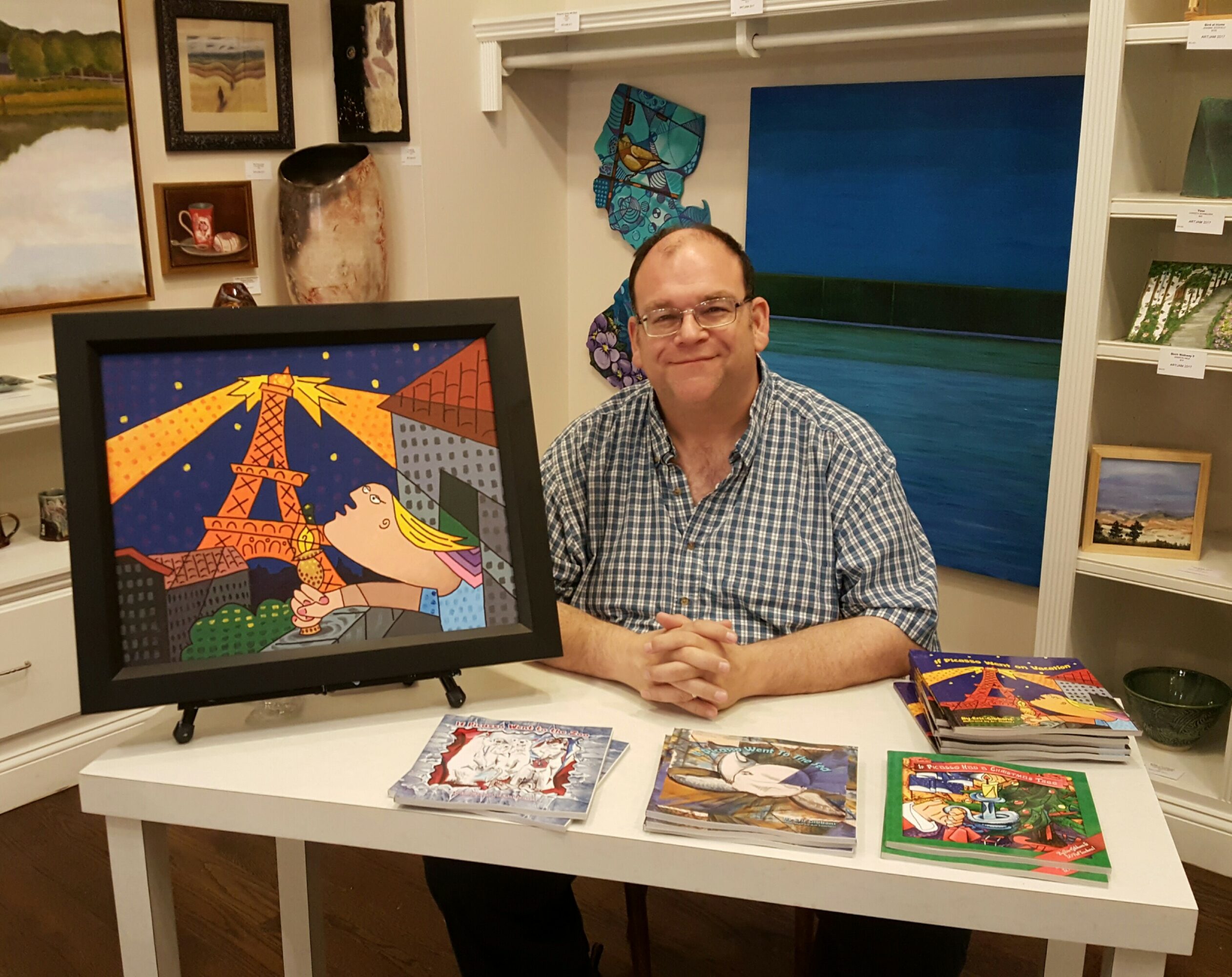 Man sitting at a table with artwork, books, and framed pictures in background, smiling at camera.