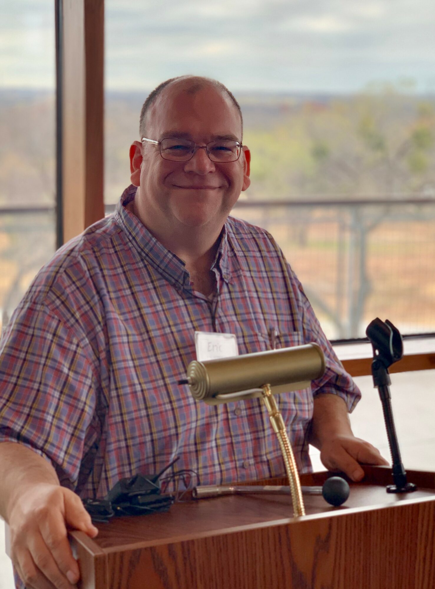 Smiling man with glasses in a plaid shirt sitting at a table with a microphone and a window background.