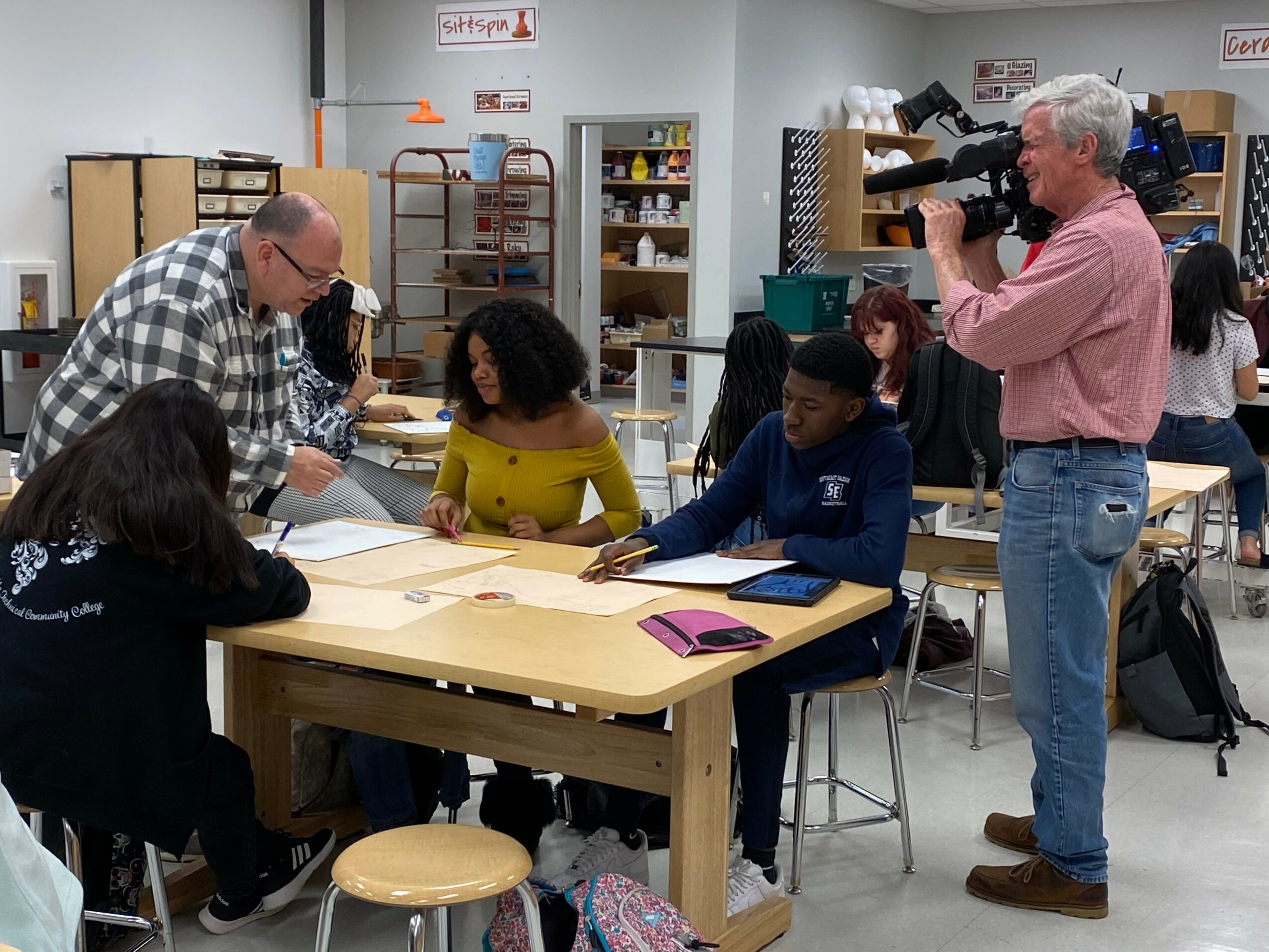 A teacher and students sit at a table in a classroom, with a cameraman filming them. Classroom shelves and students in background.