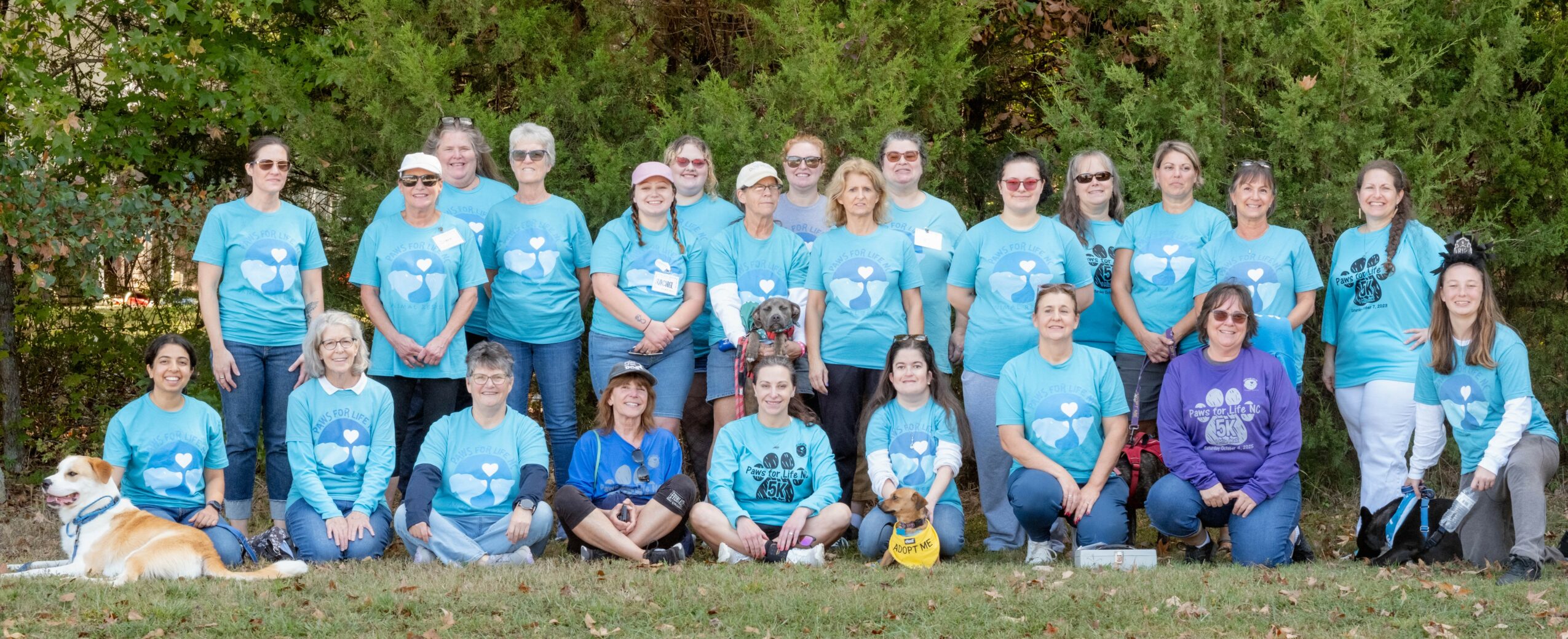 Group of people outdoors, standing and sitting, wearing matching blue shirts, with trees in the background.