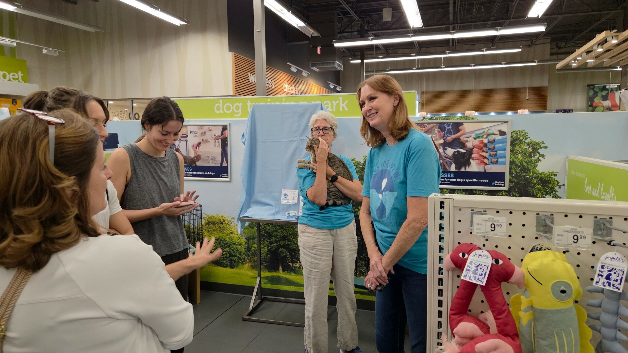 Group of women talking at an indoor event, with display boards and plush toys nearby.