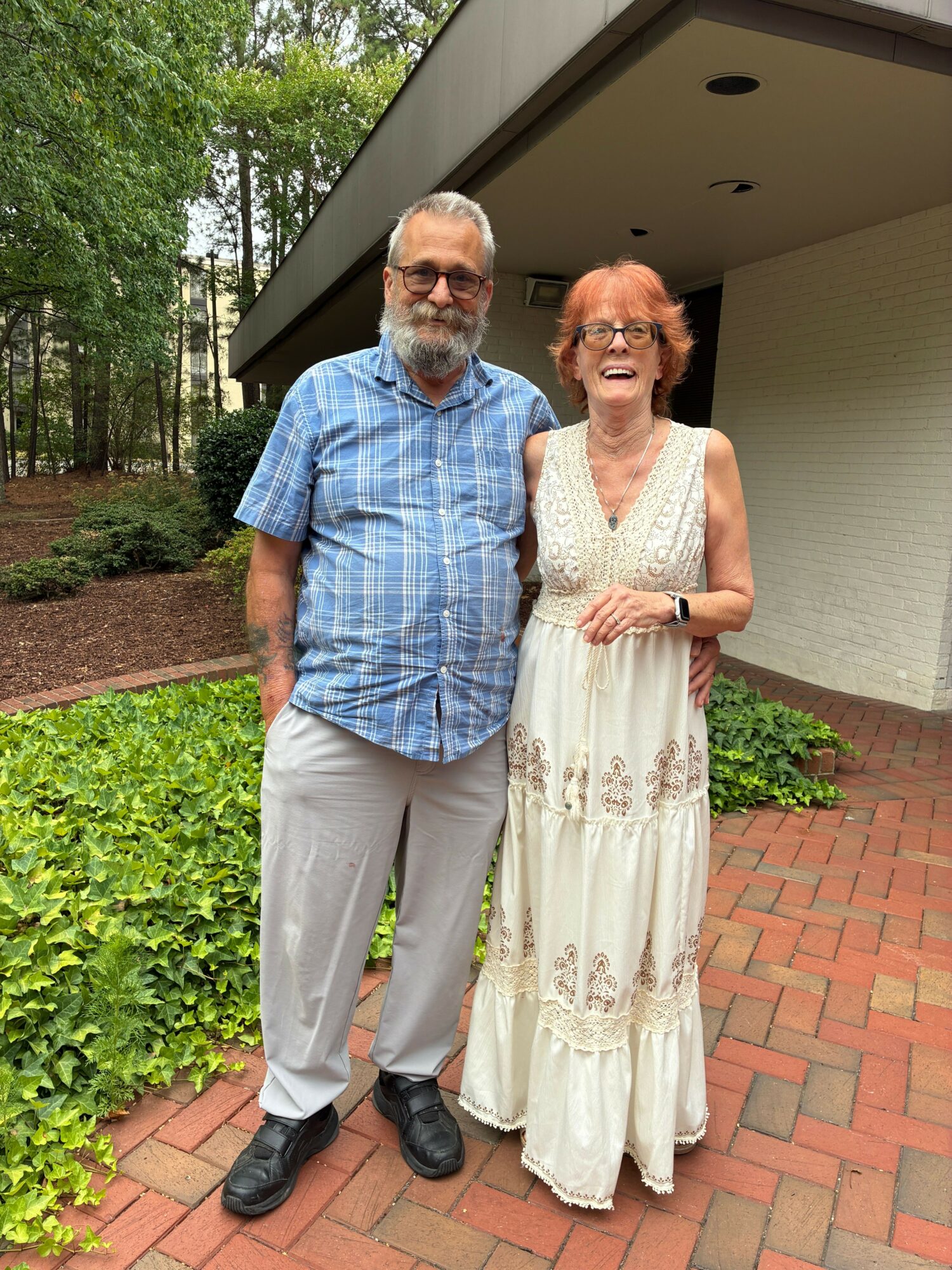 A woman in a white dress and a man in a blue shirt and gray pants stand outdoors on a brick pathway with greenery around them.