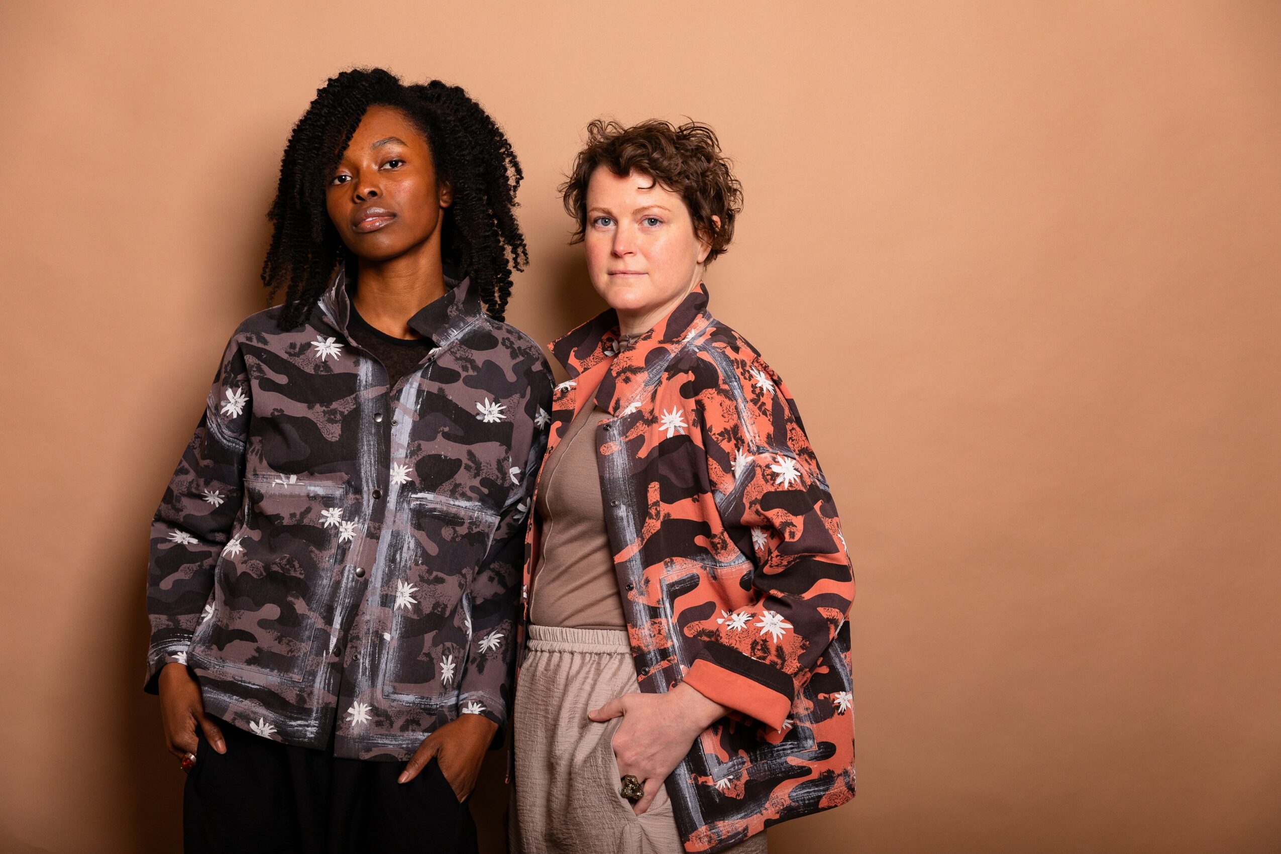 Two women stand side by side against a beige background, wearing patterned jackets and casual clothing.