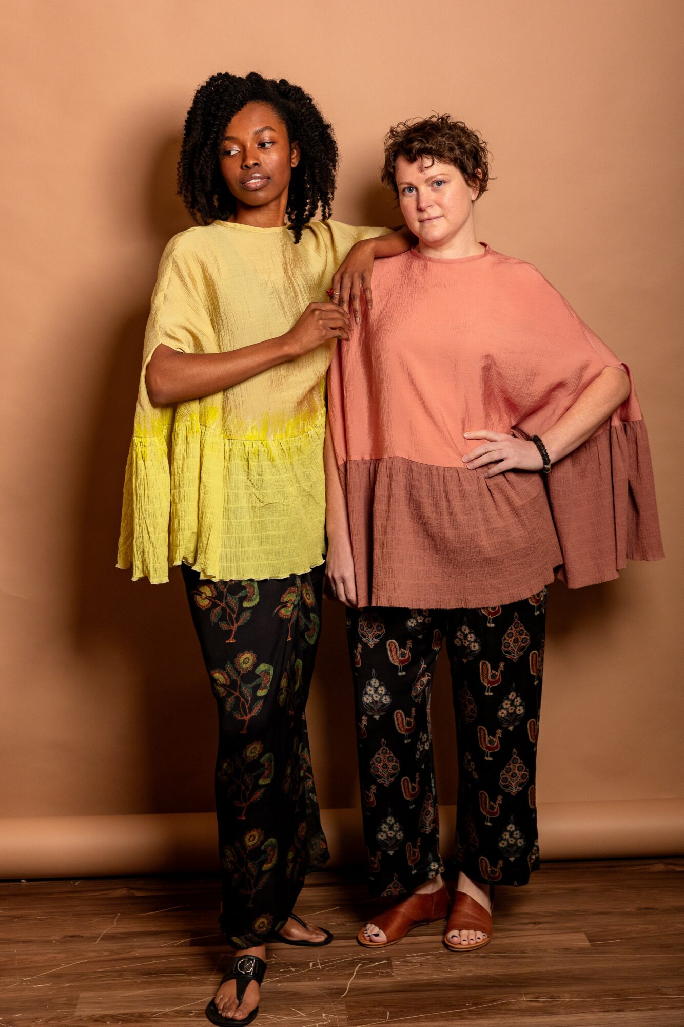 Two women stand side by side against a beige background, one with curly hair and the other with short hair, both wearing colorful, loose-fitting clothing and patterned pants.
