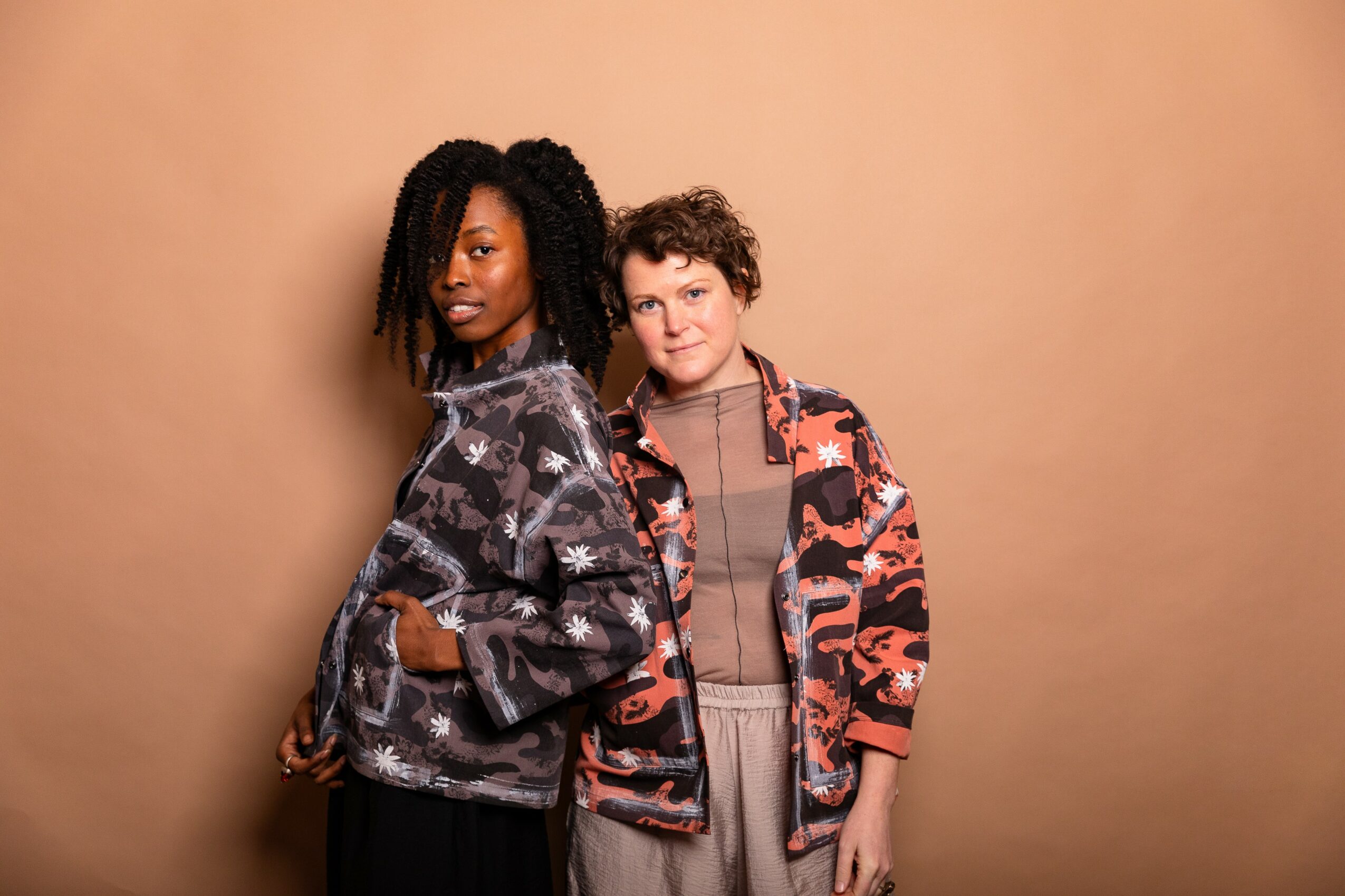 Two people standing against a plain beige background, one with long curly hair and the other with short curly hair, both wearing patterned jackets.