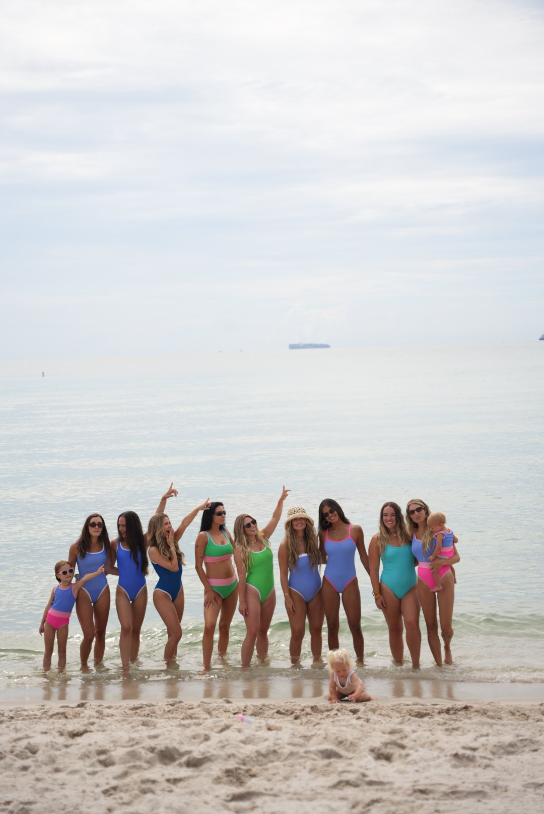 Group of women and children standing on a beach near the water, some waving, with a cloudy sky and distant ship in background.