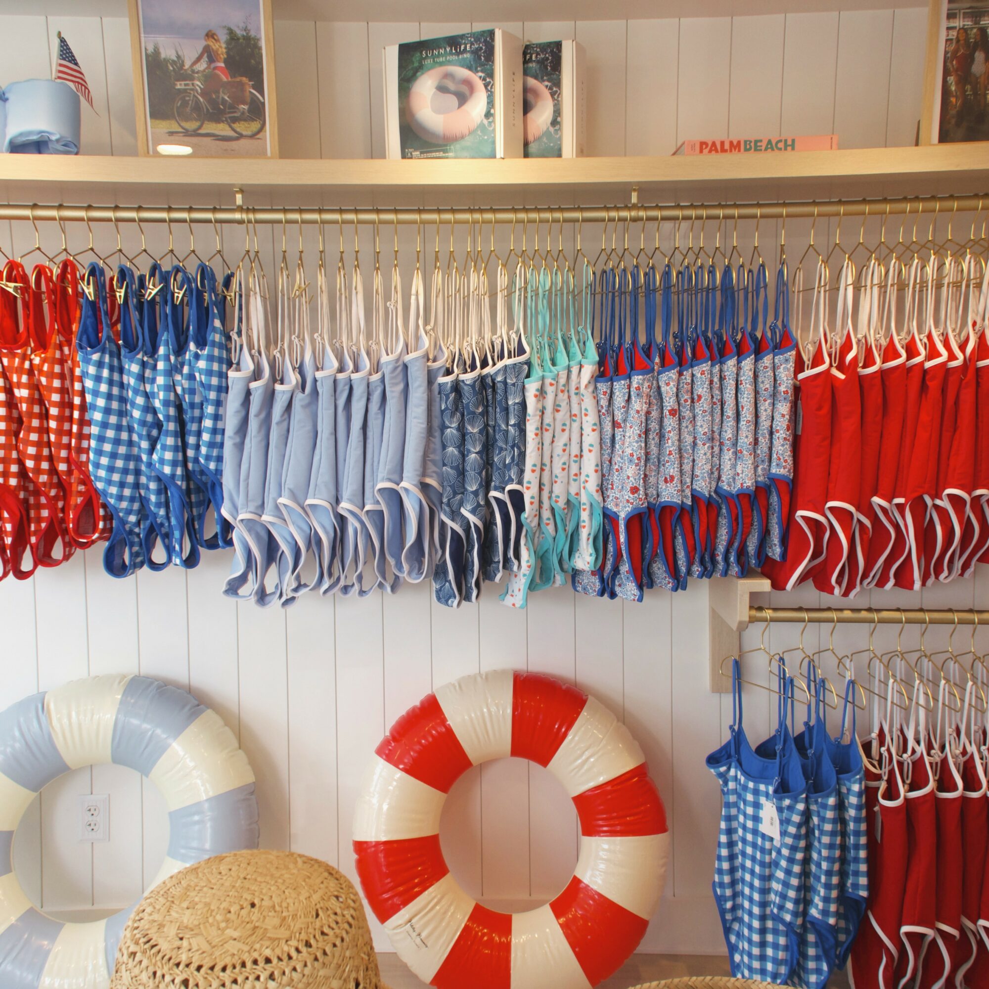 Display of colorful swimsuits hanging on a rack, with life preservers below and framed pictures above, in a retail store.