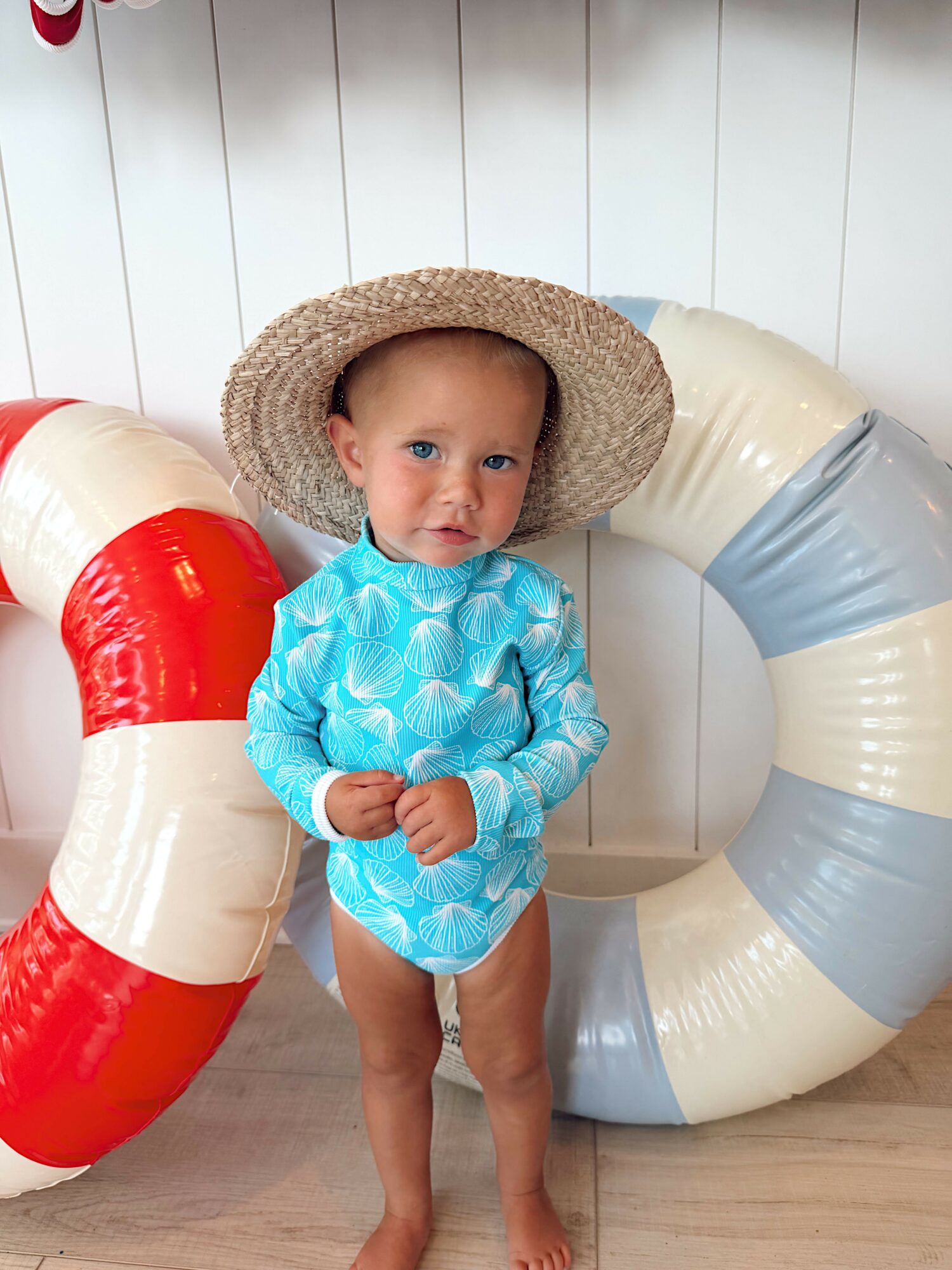 Child wearing a blue outfit and straw hat standing next to a large red and white inflatable ring, with a blue and white ring behind.