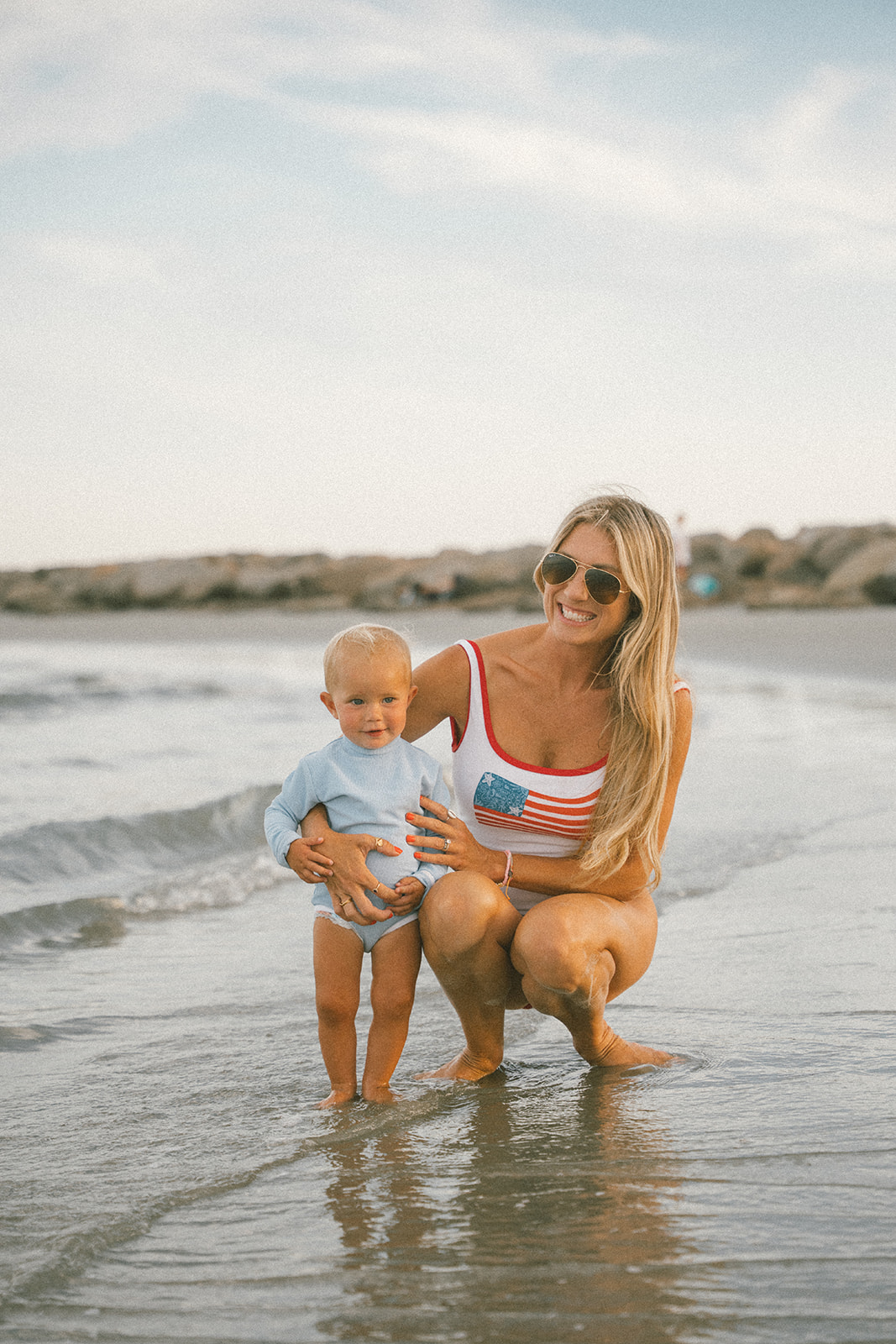 Woman and child standing in shallow ocean water at the beach, smiling, woman wearing sunglasses and a tank top with an American flag, child in light blue shirt and shorts