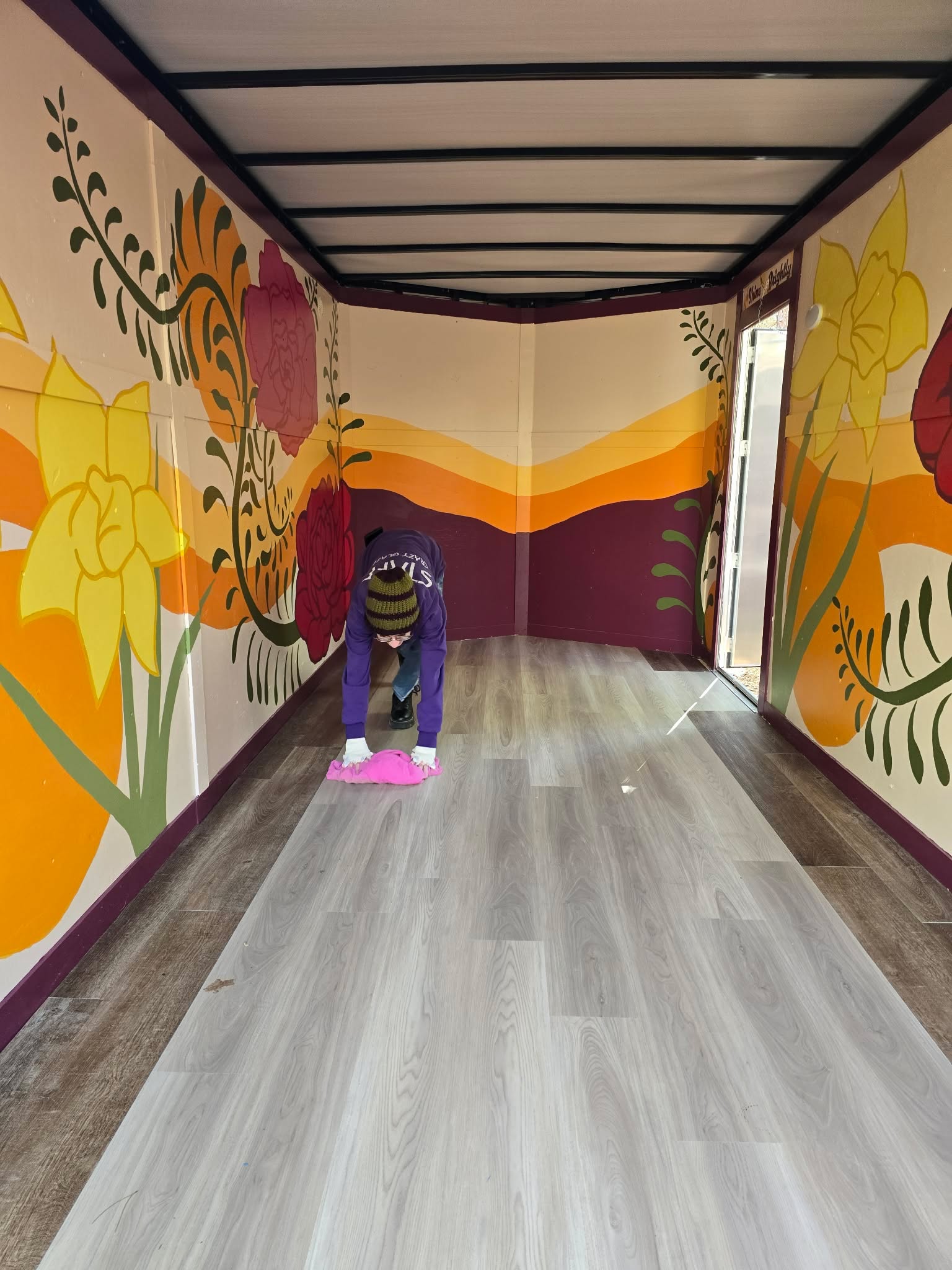 Person cleaning floor with a pink cloth in a colorful, mural-painted hallway with floral designs and a wooden floor.