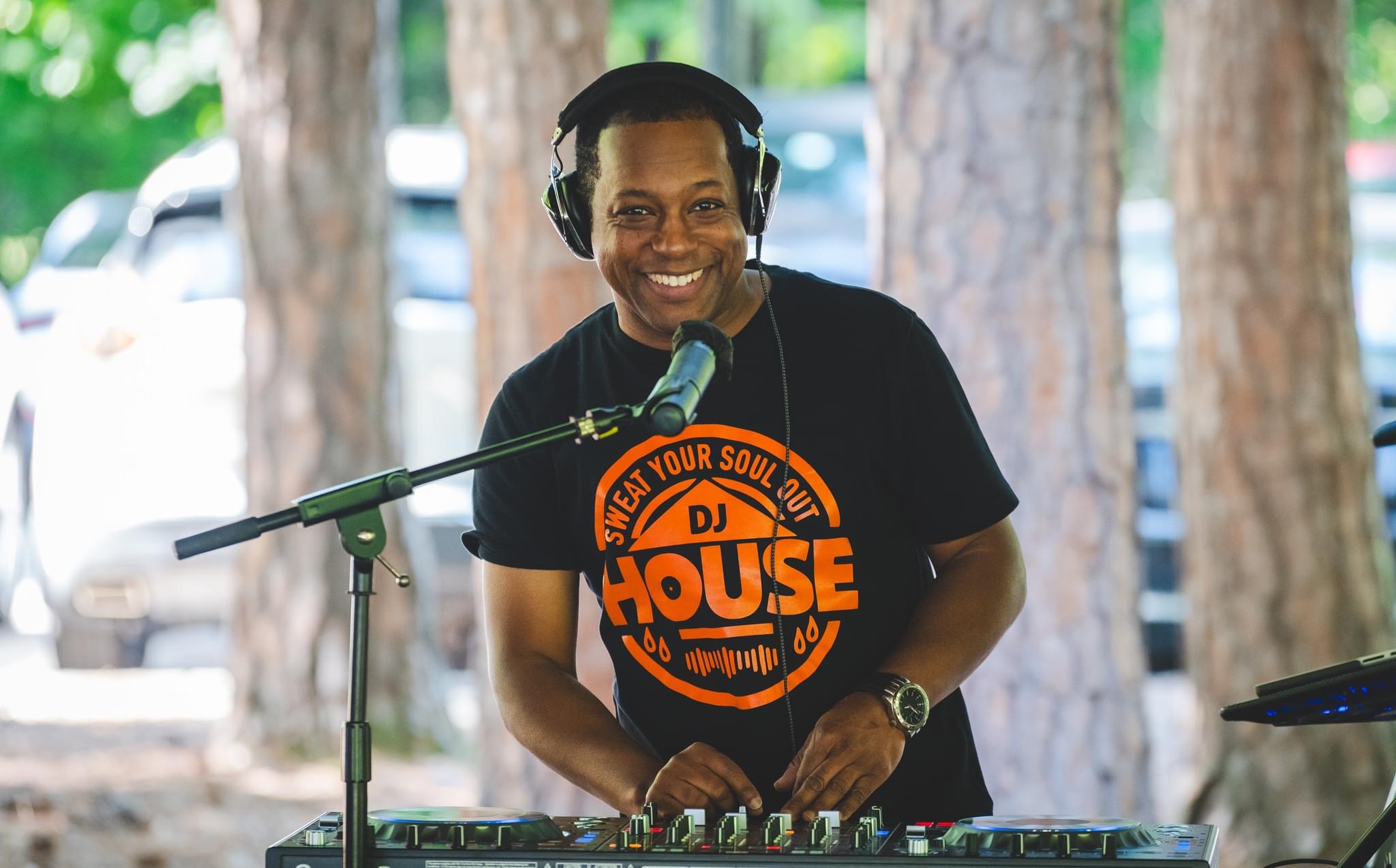 Person DJing outdoors with headphones, smiling, standing behind DJ equipment, trees in background.