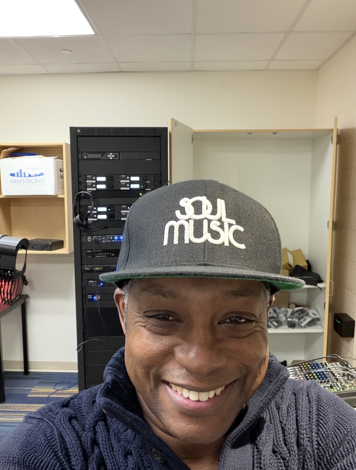 Smiling person wearing a gray cap with white text, in an indoor setting with electronic equipment and shelves.