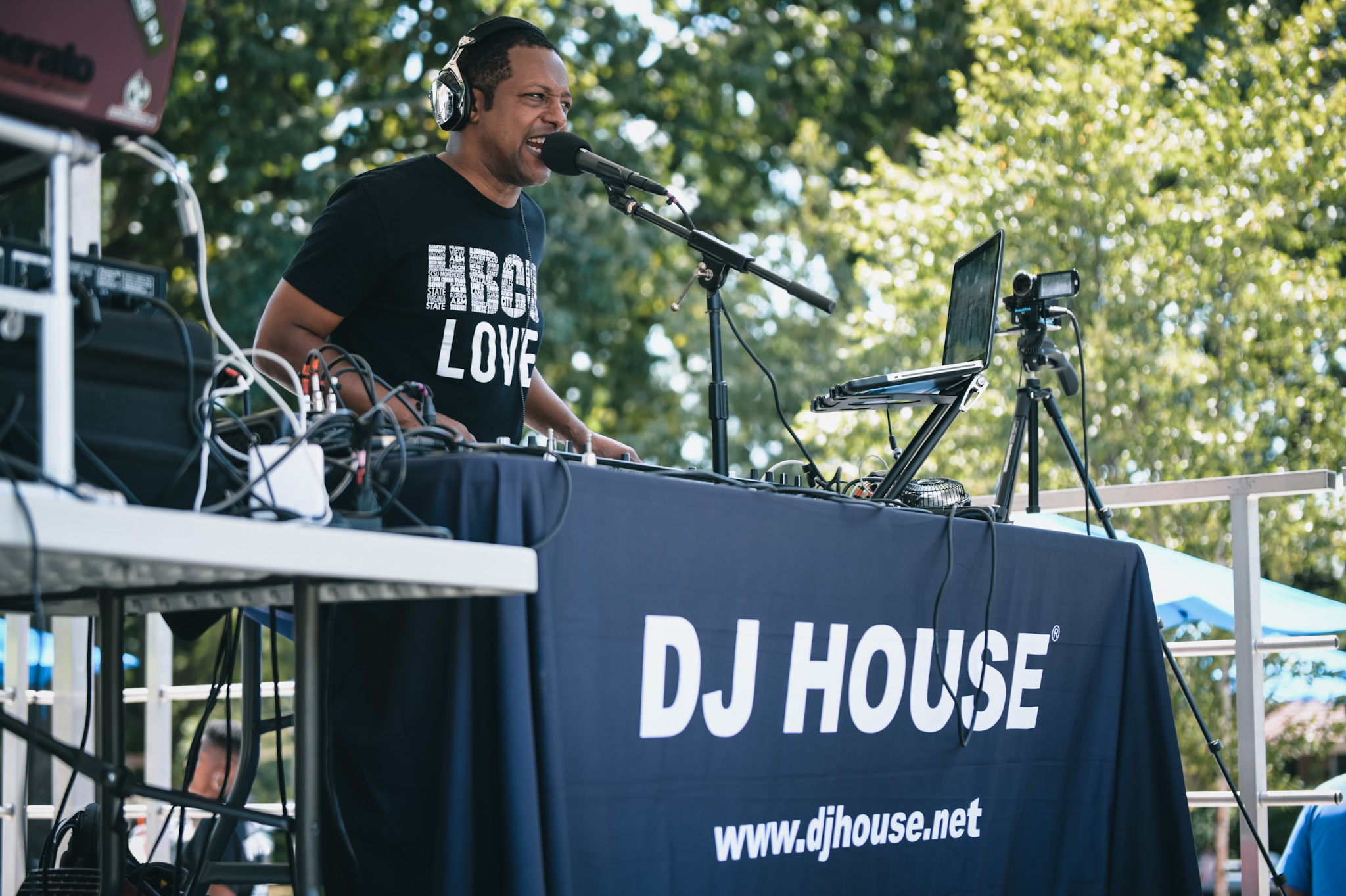 DJ performing outdoors at a table with a banner reading 'DJ HOUSE' and website, surrounded by trees and sunlight.