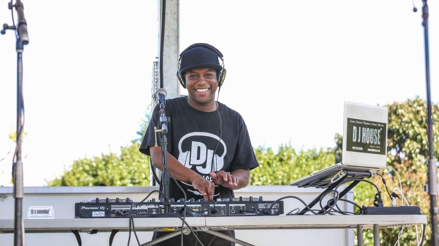 DJ smiling at a mixing console outdoors with trees in background