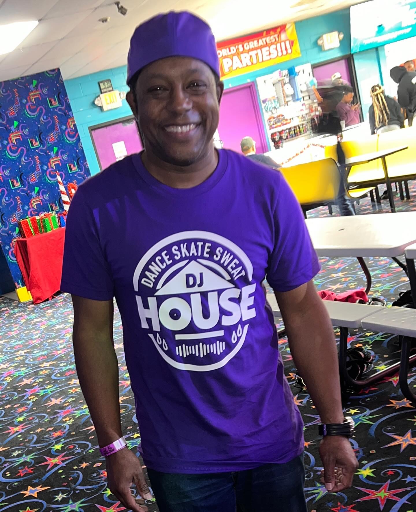 Smiling person wearing a purple hat and T-shirt with a DJ house logo, standing in an arcade or entertainment area.