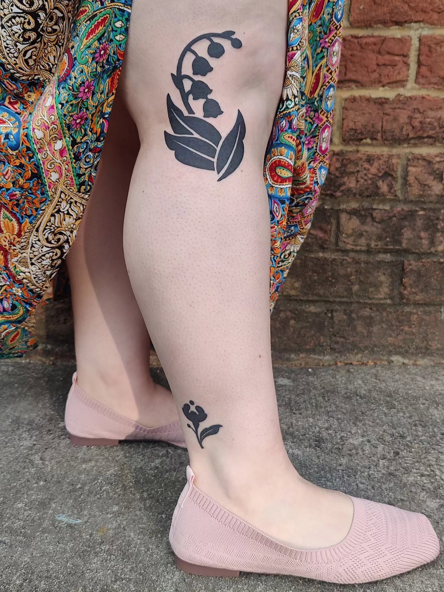 Person's leg with a black floral tattoo, wearing pink shoes, against a brick wall background.