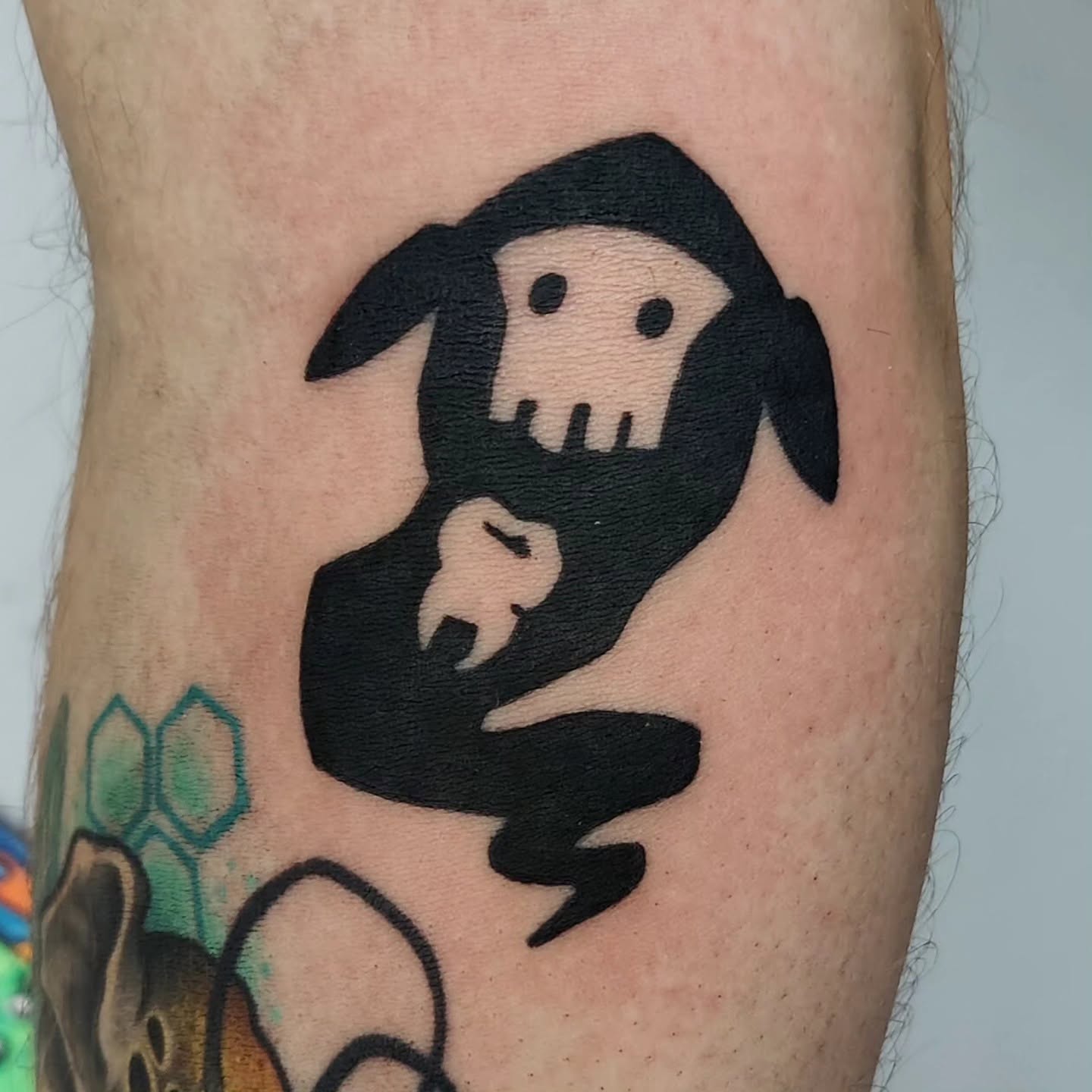 Black tattoo of a stylized creature with a skull face and floppy ears, holding a small object, on skin.