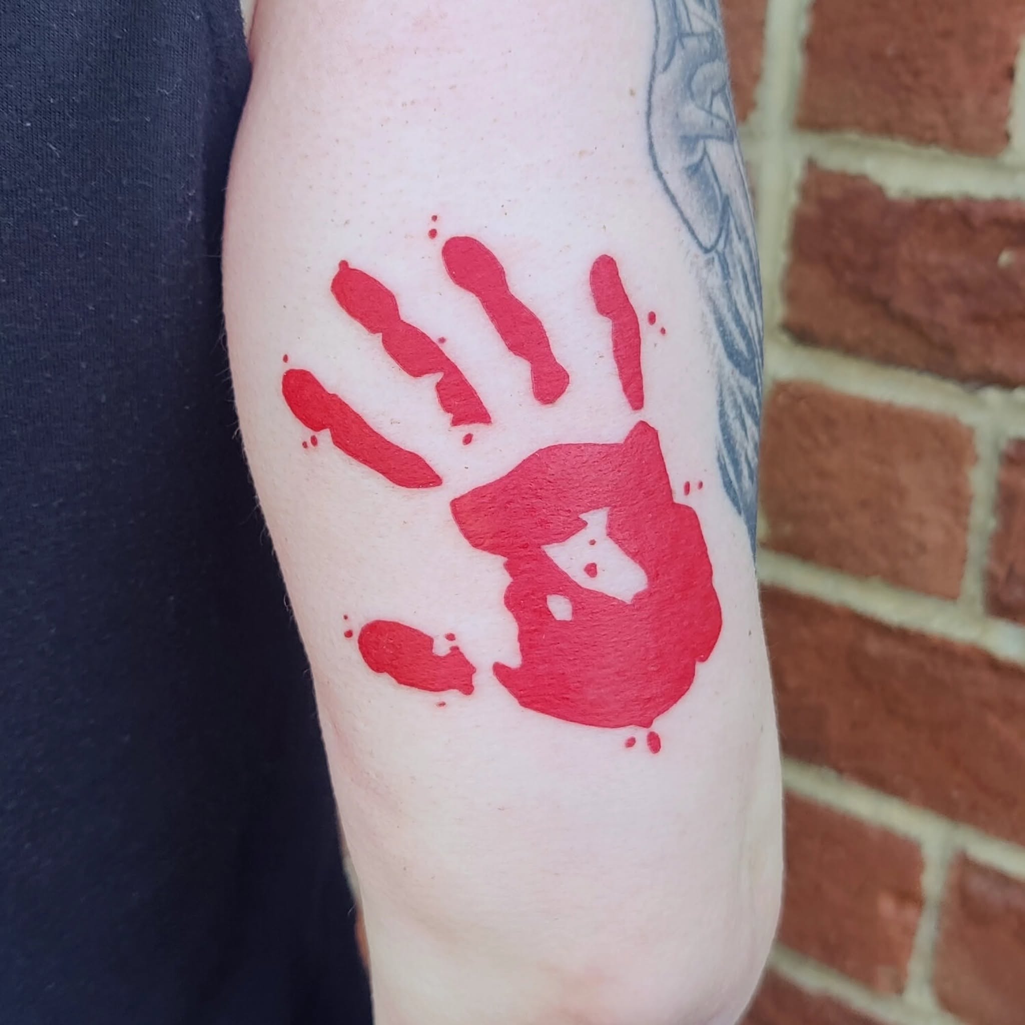 Red handprint tattoo on person's arm with a brick wall background.