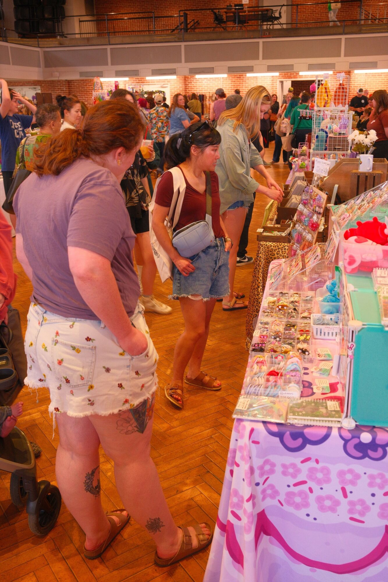 People browsing items on a table at an indoor market or event, with wooden flooring and various products displayed.