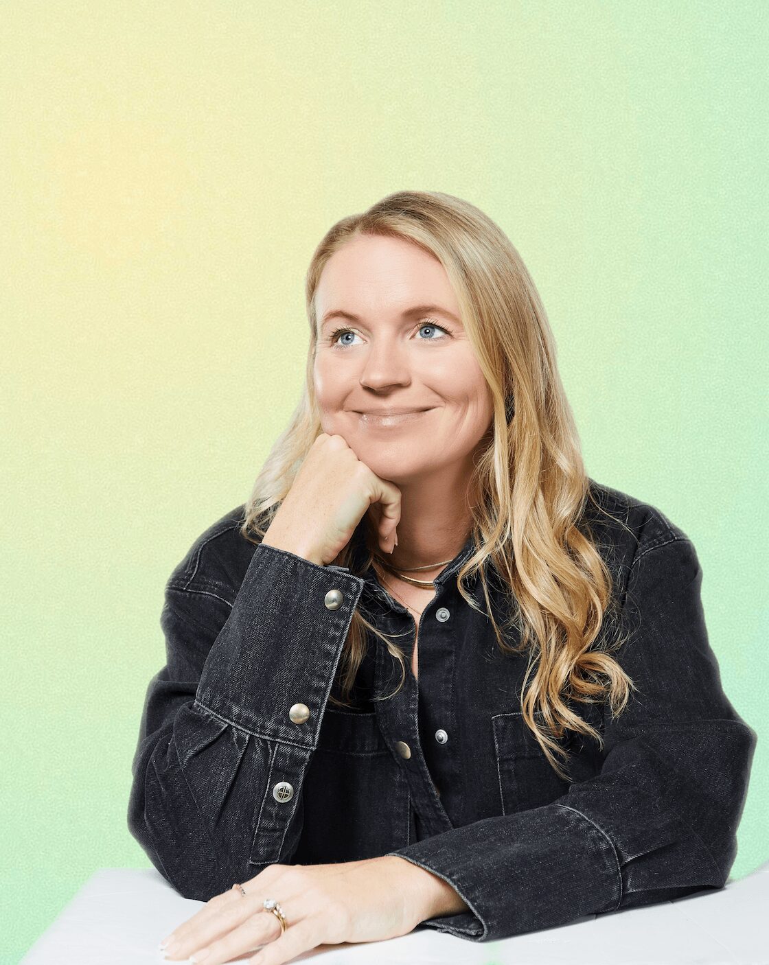 Woman with long blonde hair wearing a black denim jacket, smiling and resting her chin on her hand, against a pastel gradient background.