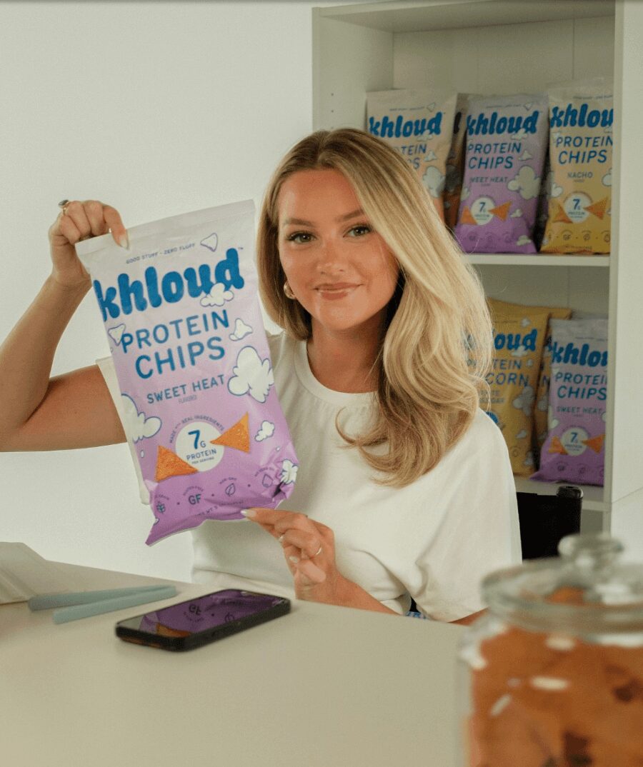 Woman with blonde hair holding a bag of protein chips, smiling, in a store with more chips on shelves behind her.